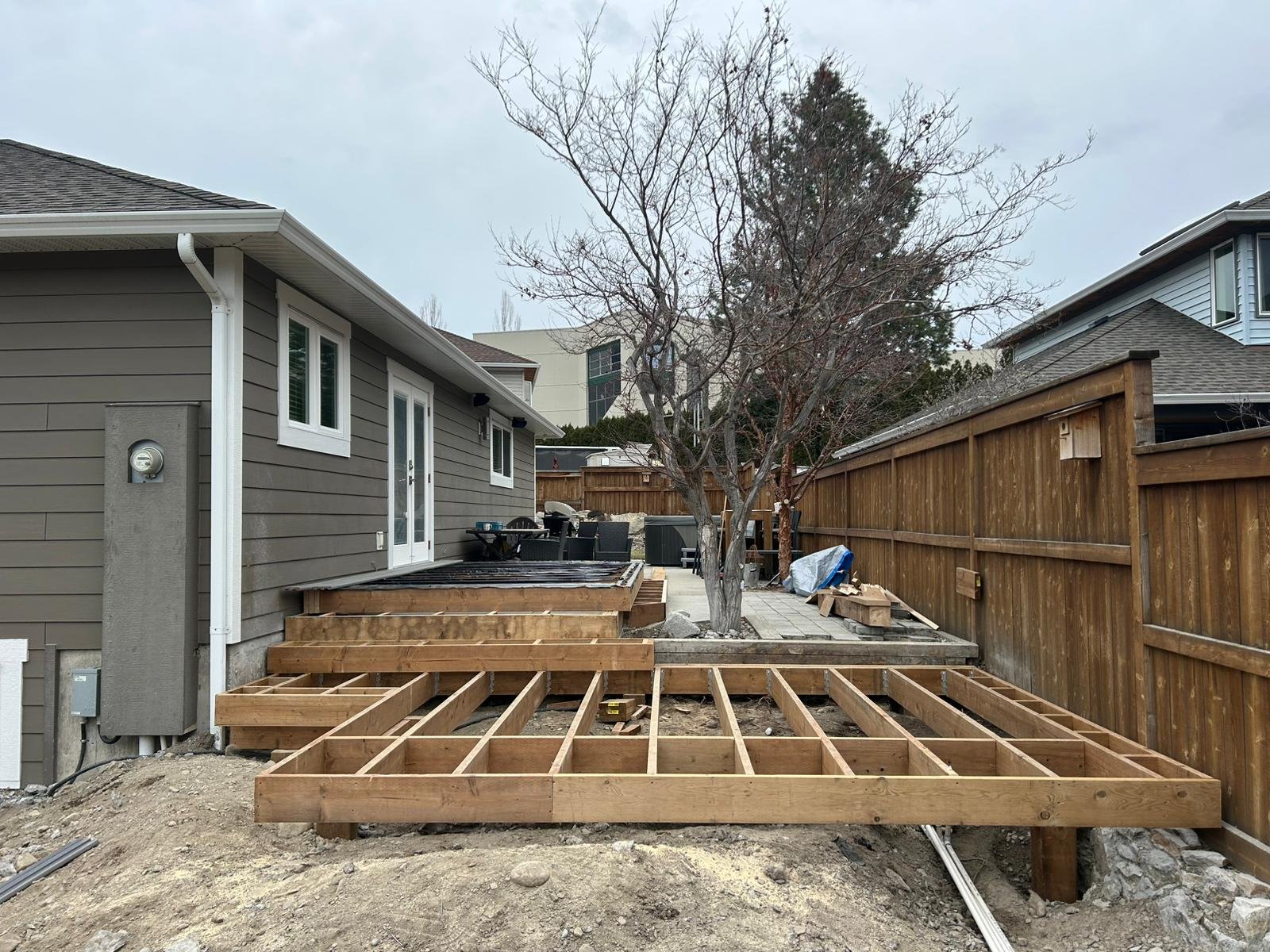 Construction of a deck or patio extension in a backyard with wooden framing, next to a gray house with white trim and a leafless tree, enclosed by a wooden fence, under an overcast sky.