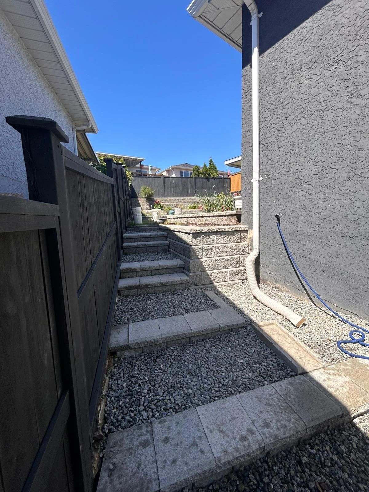 Residential backyard with stone steps leading up to a garden, a gravel pathway, a black wooden fence on the left, and a gray wall on the right, with a clear blue sky overhead.