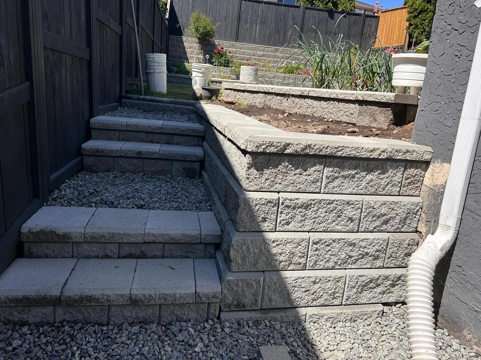 A backyard landscape featuring a raised stone planter with a gravel pathway and stone steps leading up to a garden bed with plants, flowers, and containers, enclosed by a black wooden fence.