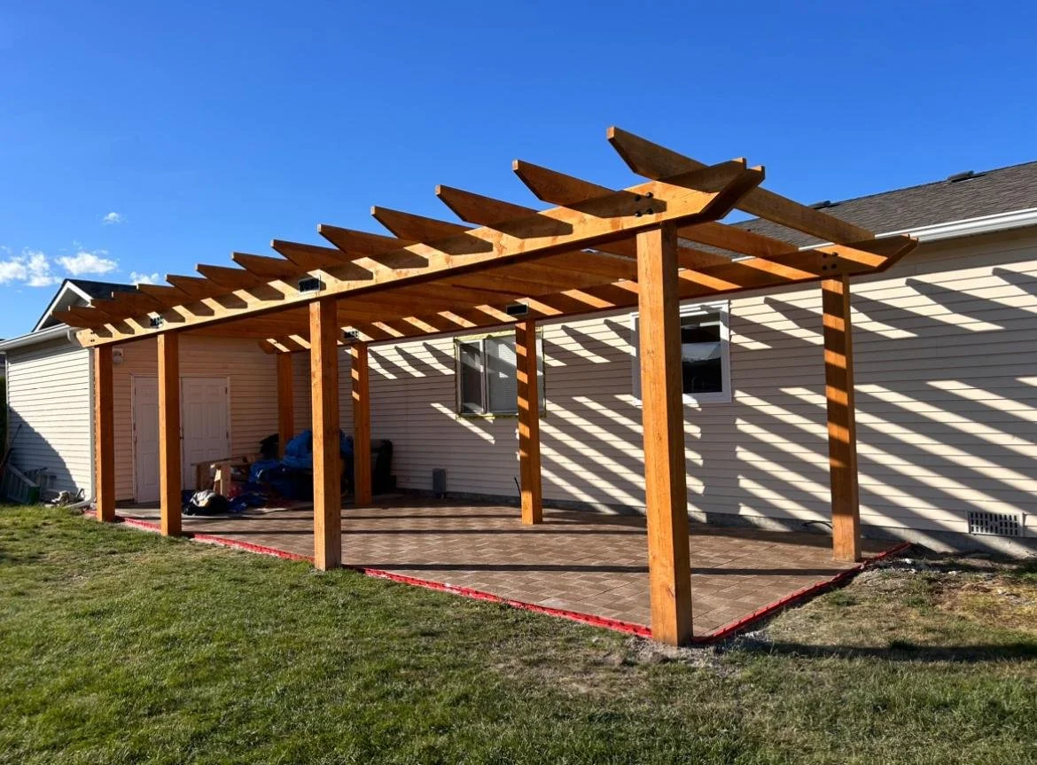 Wooden pergola under construction on a patio outside a beige house with vinyl siding, with shadow patterns cast on the wall and patio floor, on a sunny day.