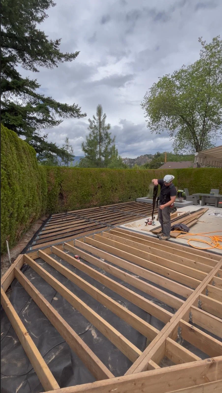Person working on building a deck frame outdoors with trees and cloudy sky in the background.