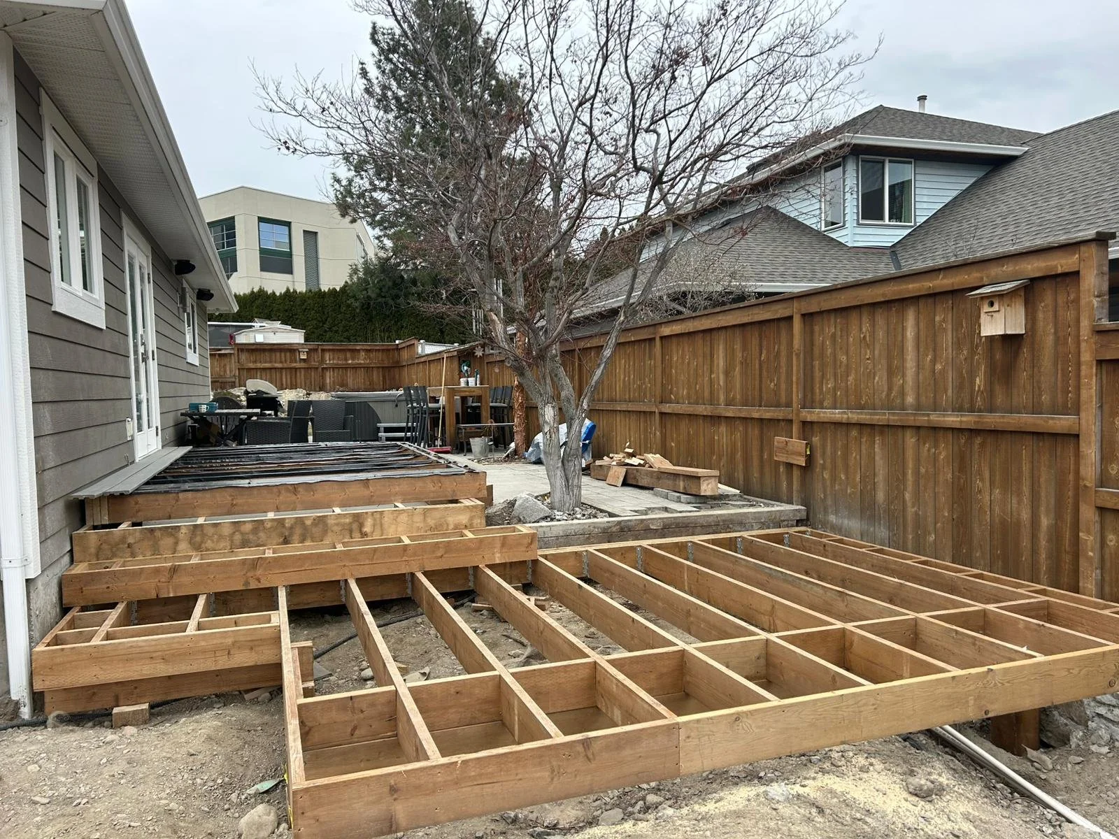 Wooden framing for a deck under construction in a backyard, with a tree and neighboring houses in the background.