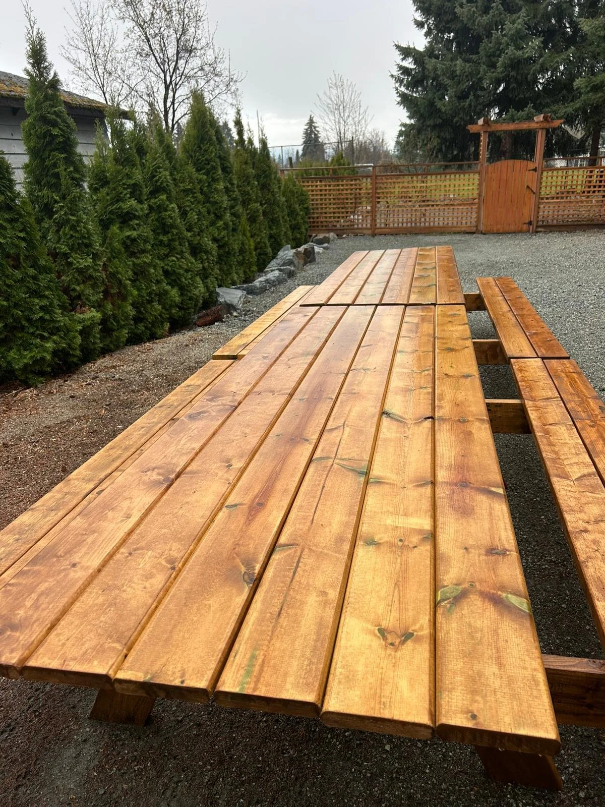 A newly made wooden picnic table with attached benches, set outdoors on a gravel surface, with green shrubs and a fence in the background.