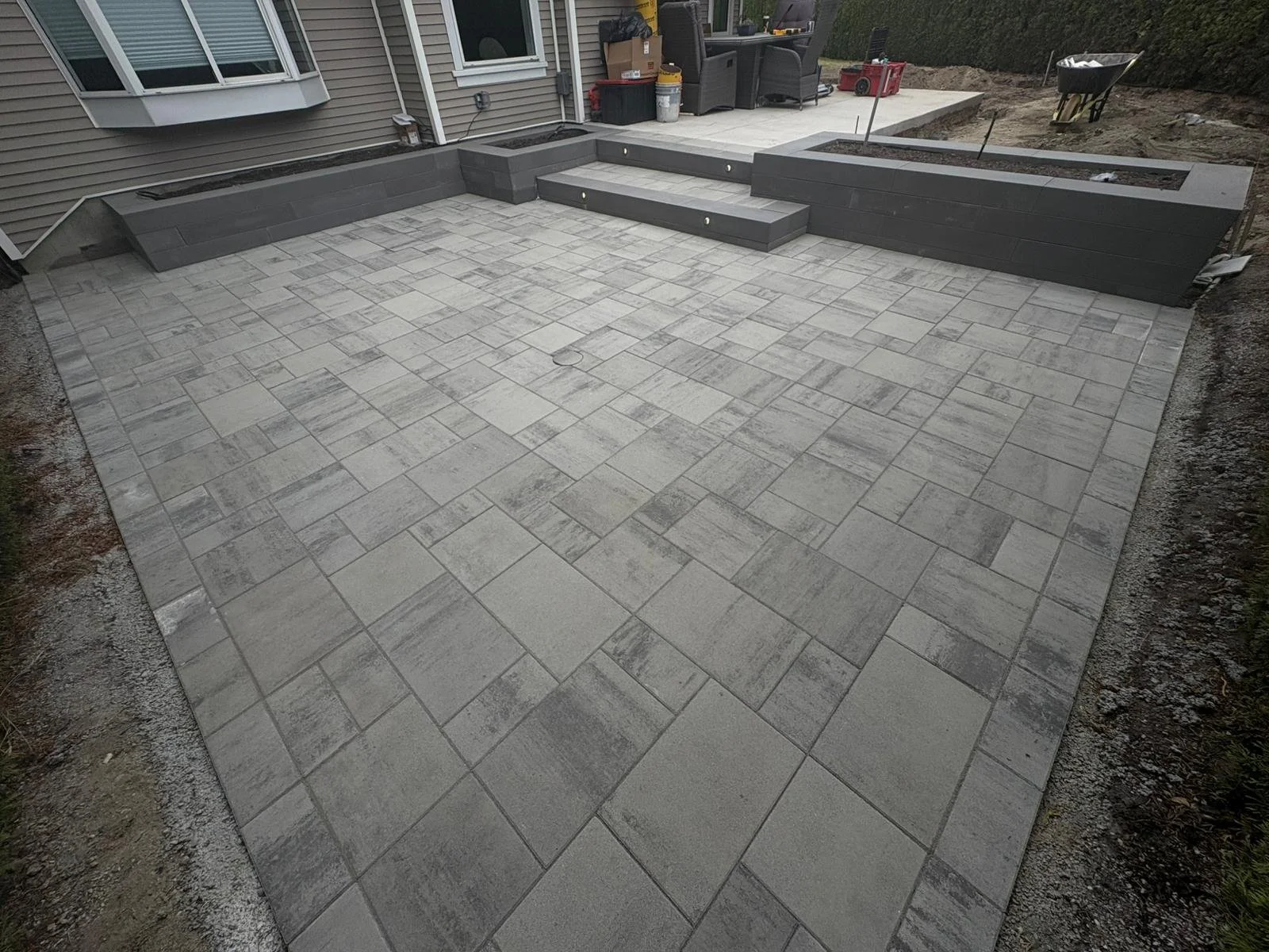 A freshly completed concrete patio with decorative paving stones, surrounded by planter beds and a house with exterior siding, outdoor furniture, a wheelbarrow, and construction materials.