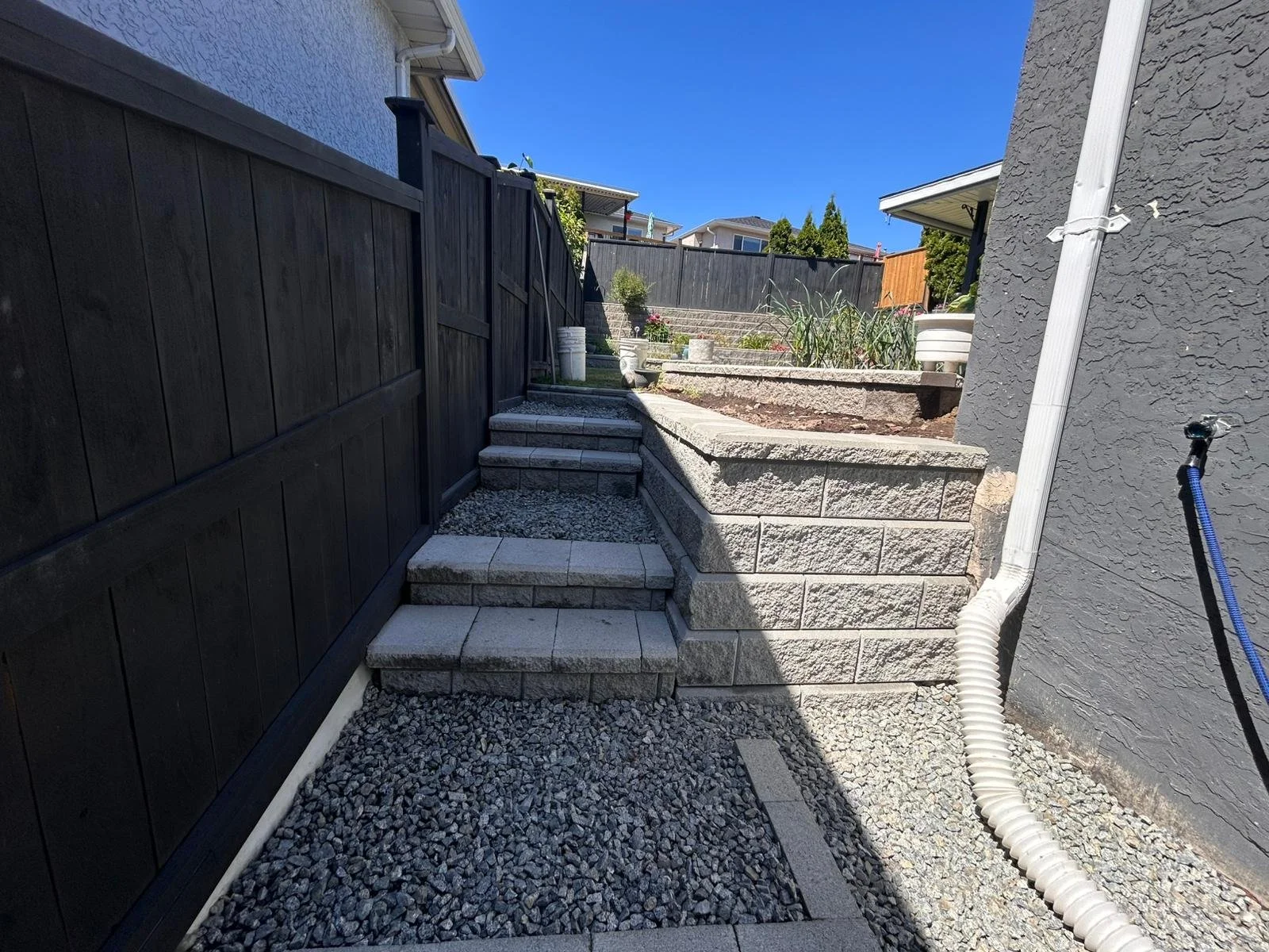 A backyard with stone stairs leading up to a garden area with potted plants and a tiered stone retaining wall. A dark wooden fence lines the left side, and a gray house wall is on the right. The sky is clear and blue.