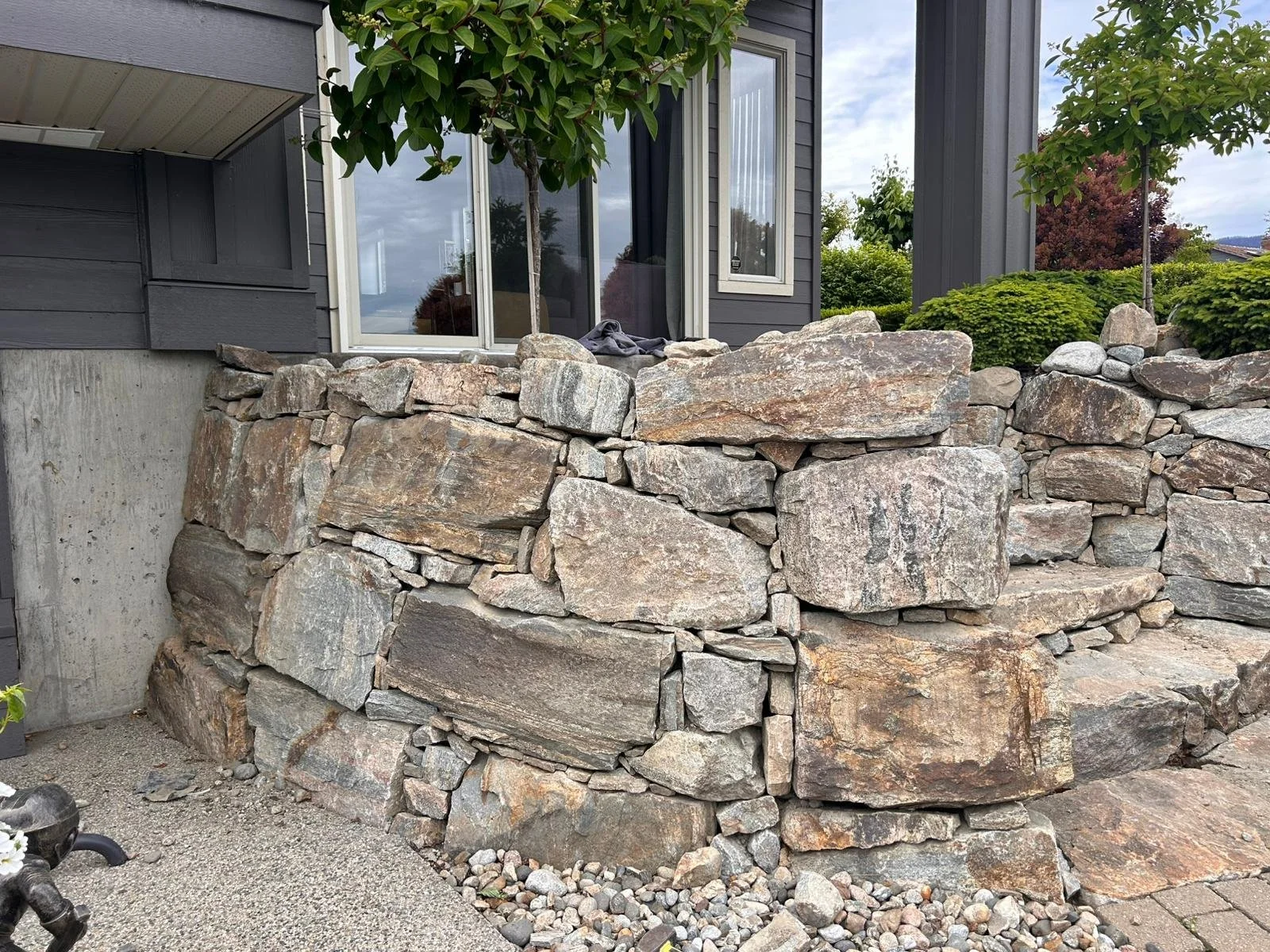 A stone retaining wall made of large, irregularly shaped rocks in front of a house with dark siding and glass sliding doors, surrounded by landscaping with small trees and bushes.