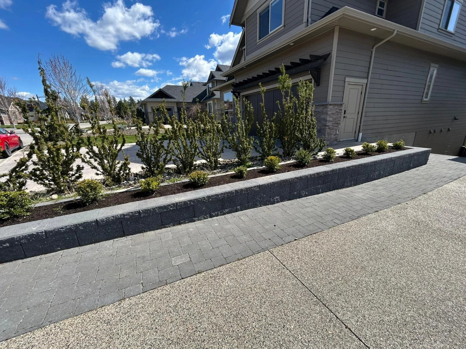 A landscaped front yard with a raised flower bed containing small trees and shrubs, with a sidewalk and a gray house in the background under a partly cloudy sky.