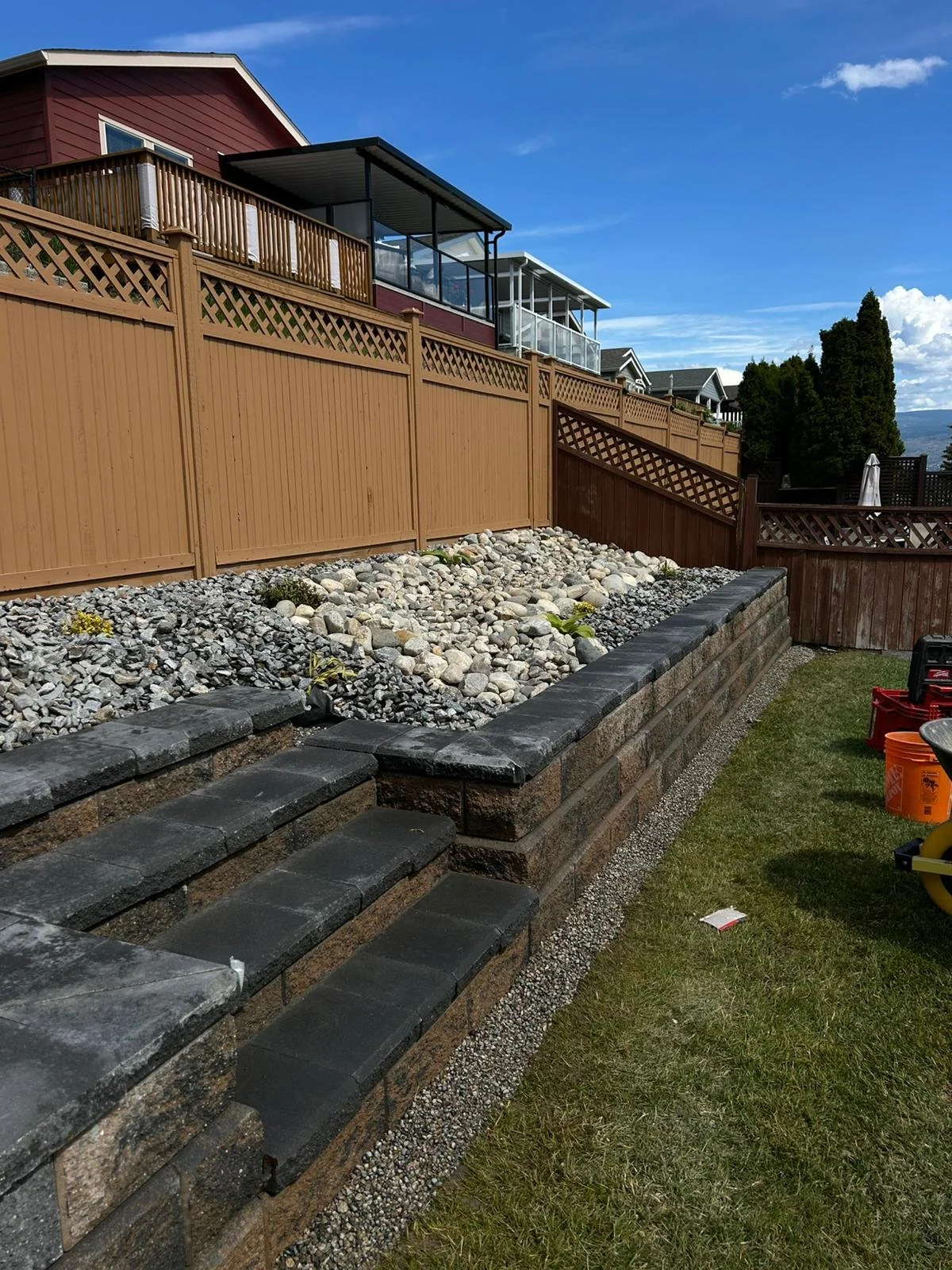 Backyard with a retaining wall made of bricks and stones, with wooden stairs, a lawn, and a privacy fence, and houses with balconies and a blue sky in the background.