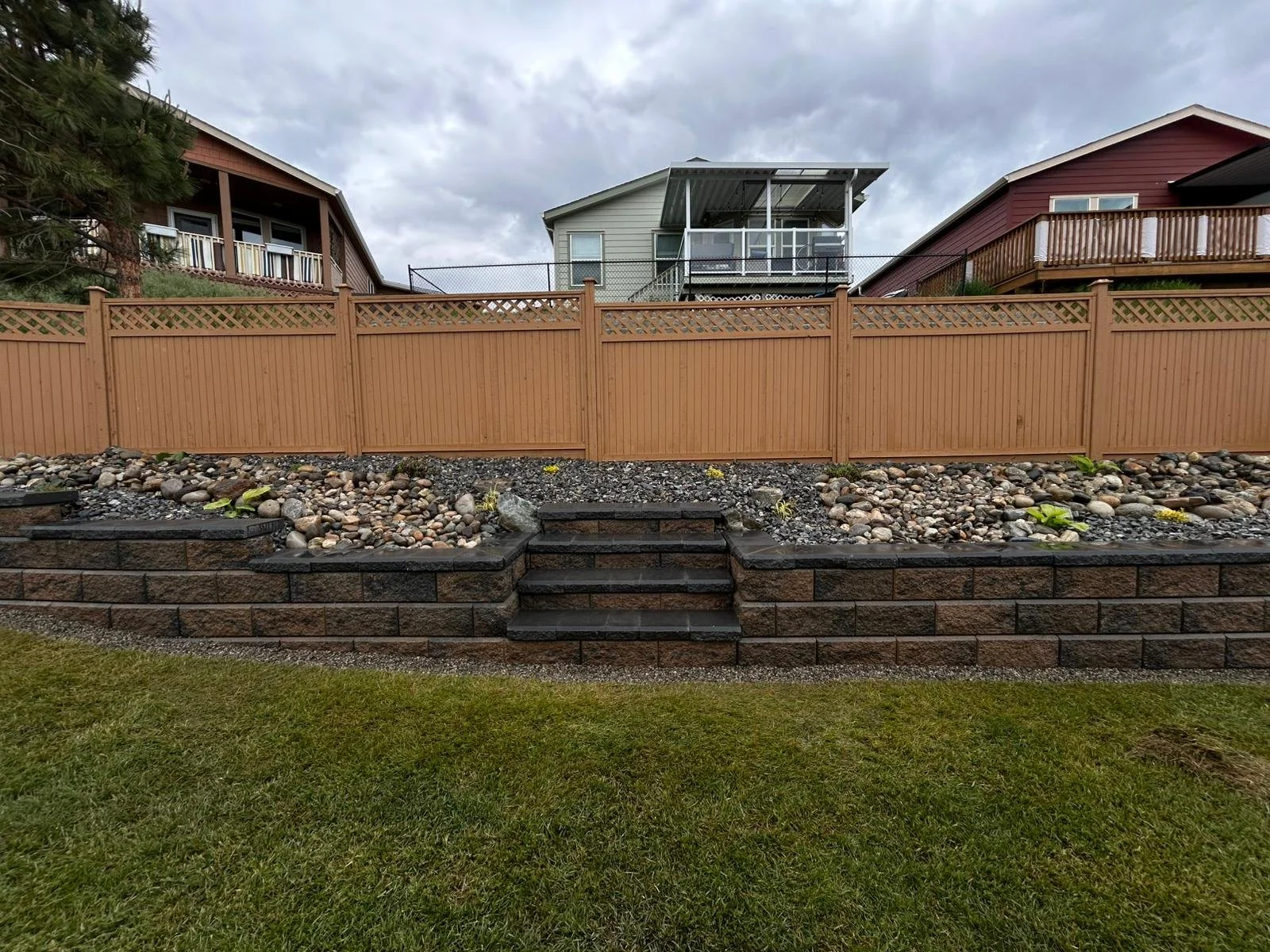 Backyard with stone retaining wall, small stairs, and fenced yard, with neighboring houses and cloudy sky visible.