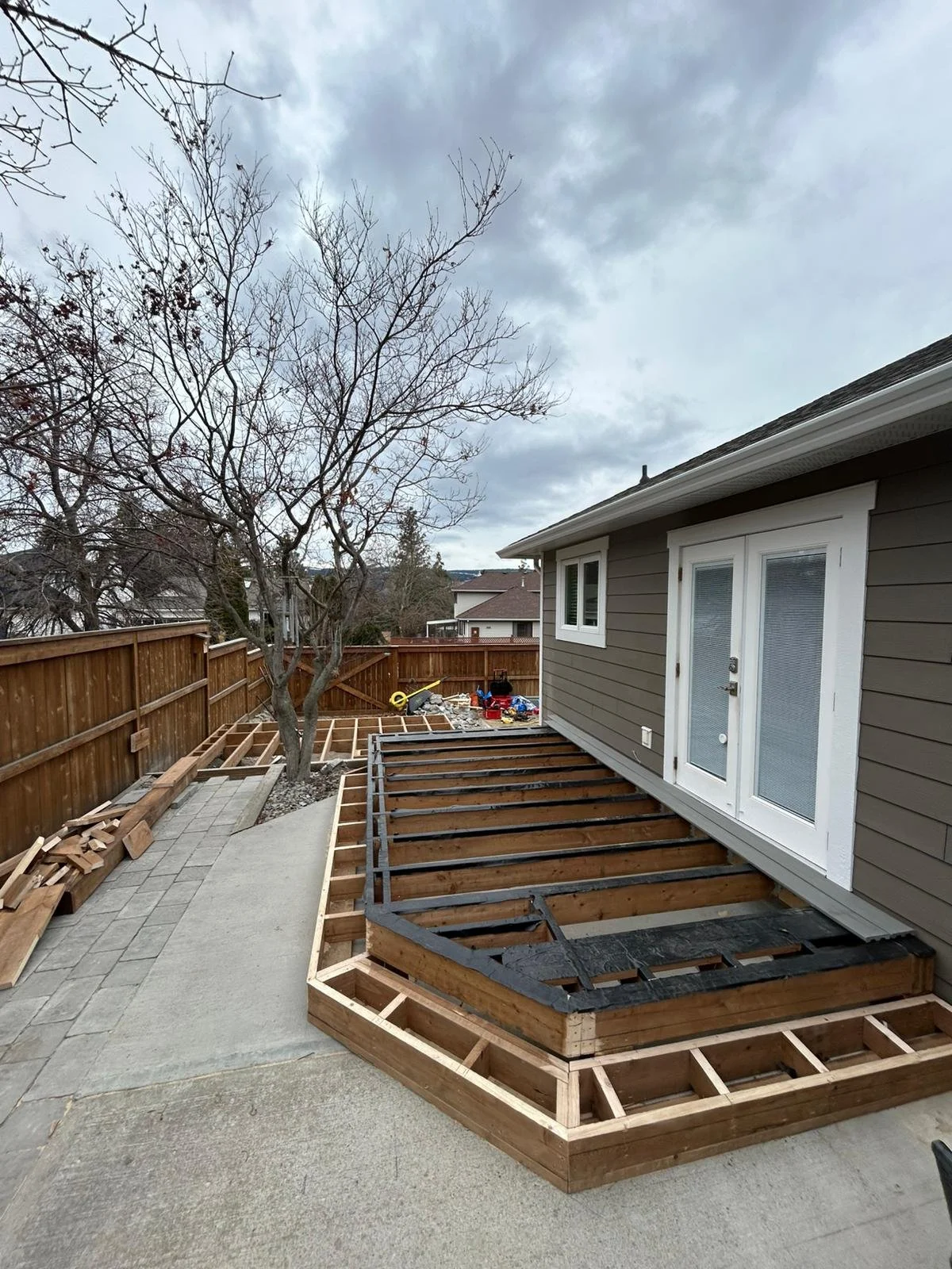 Backyard construction of a deck showing a partially built wooden frame with stairs, next to a house with gray siding and white trim, while a tree without leaves and construction tools are visible in the background.