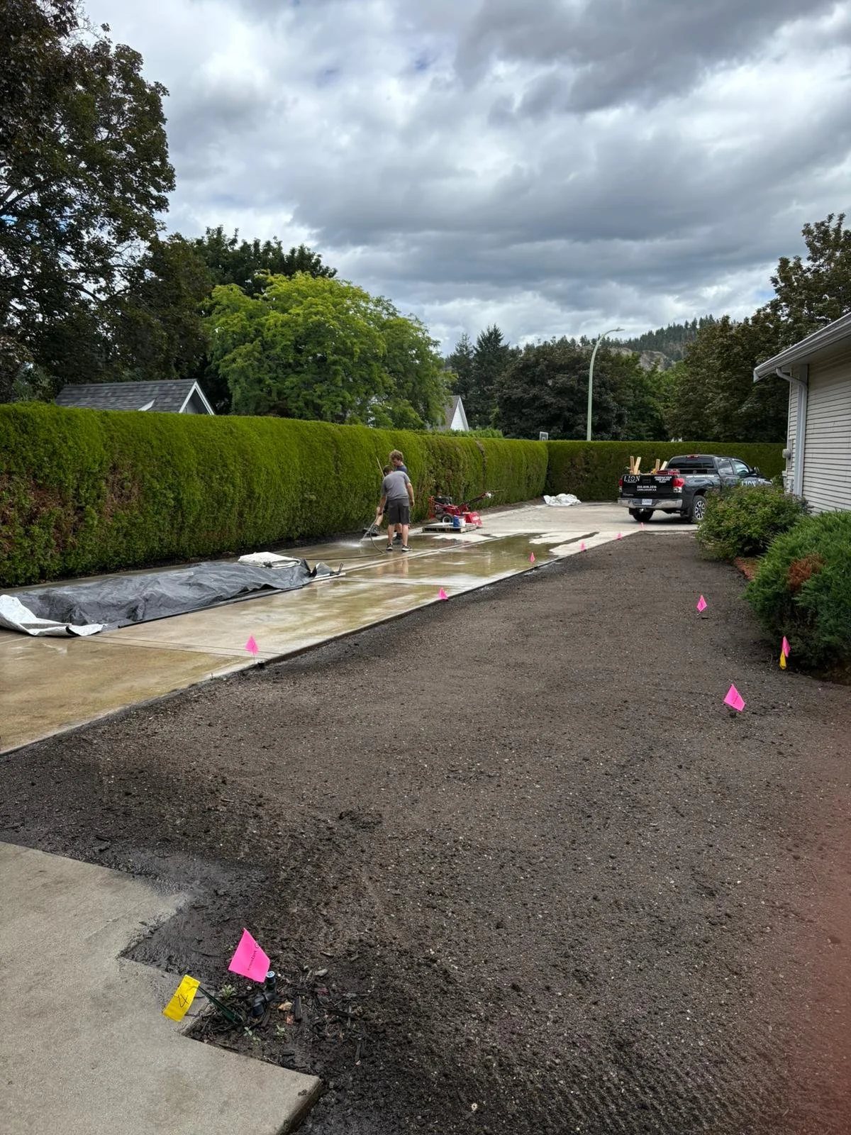 People laying down fresh concrete on a driveway, with orange flags marking the edges, a black truck parked nearby, and a lush green hedge and trees in the background under cloudy skies.