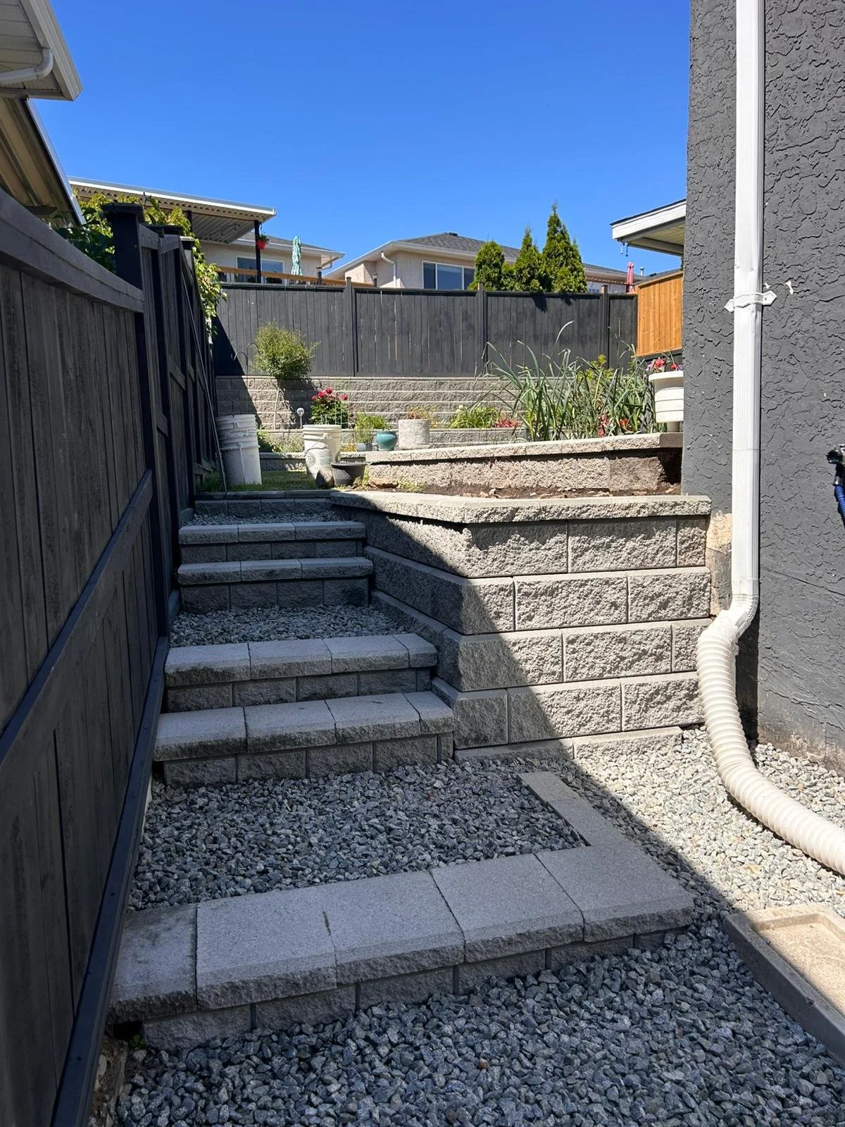 Backyard with stone stairs leading up to a garden bed with plants, bordered by a high wooden fence and a gray house wall, under a clear blue sky.