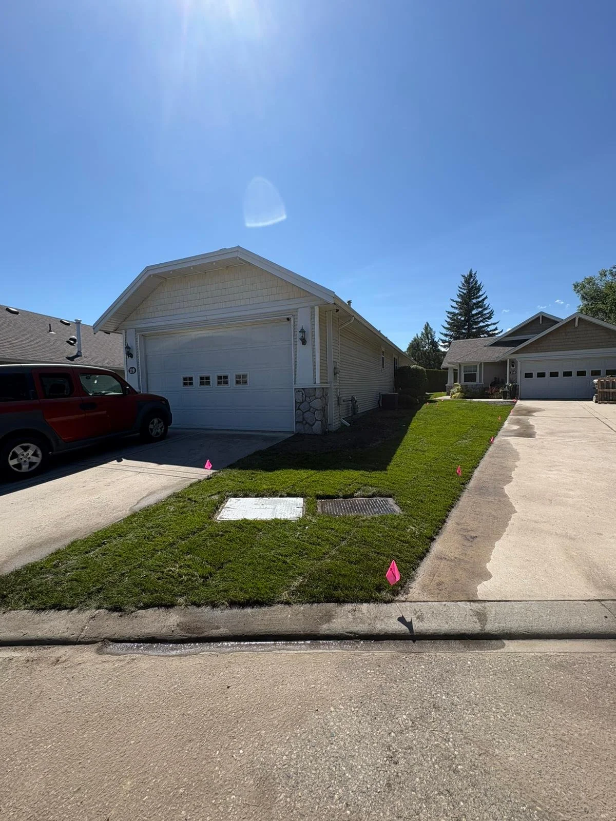 Residential driveway with grass strip in front of two detached garages, a red SUV parked on the left, and pink flags marking areas near the sidewalk and grass.