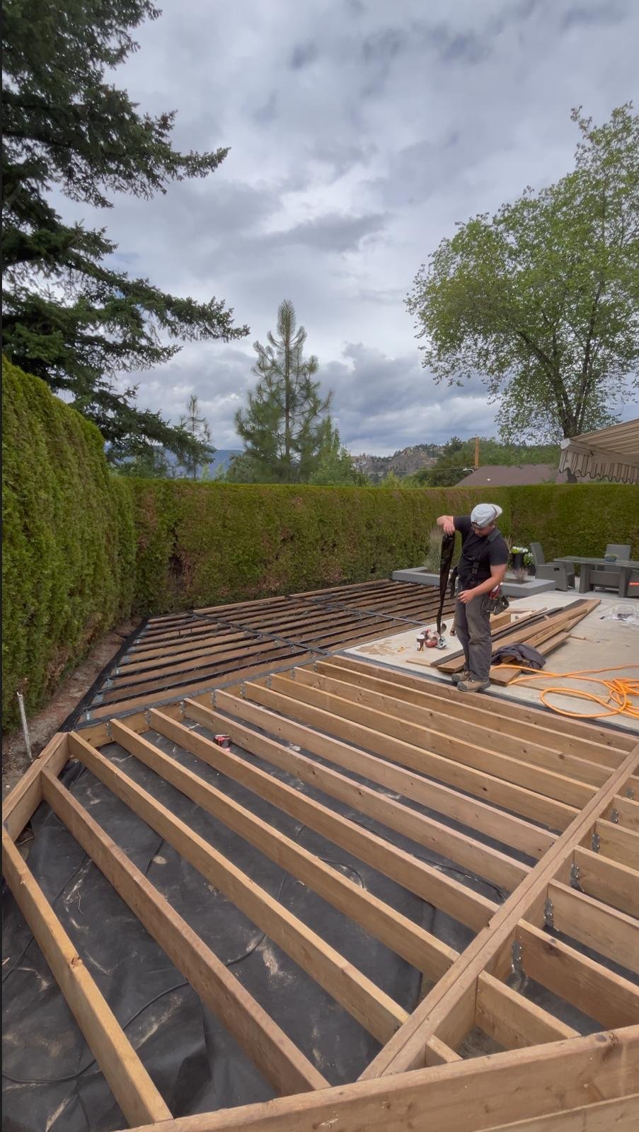 A person working on a wooden deck frame outdoors during cloudy weather, surrounded by trees and a hedge.