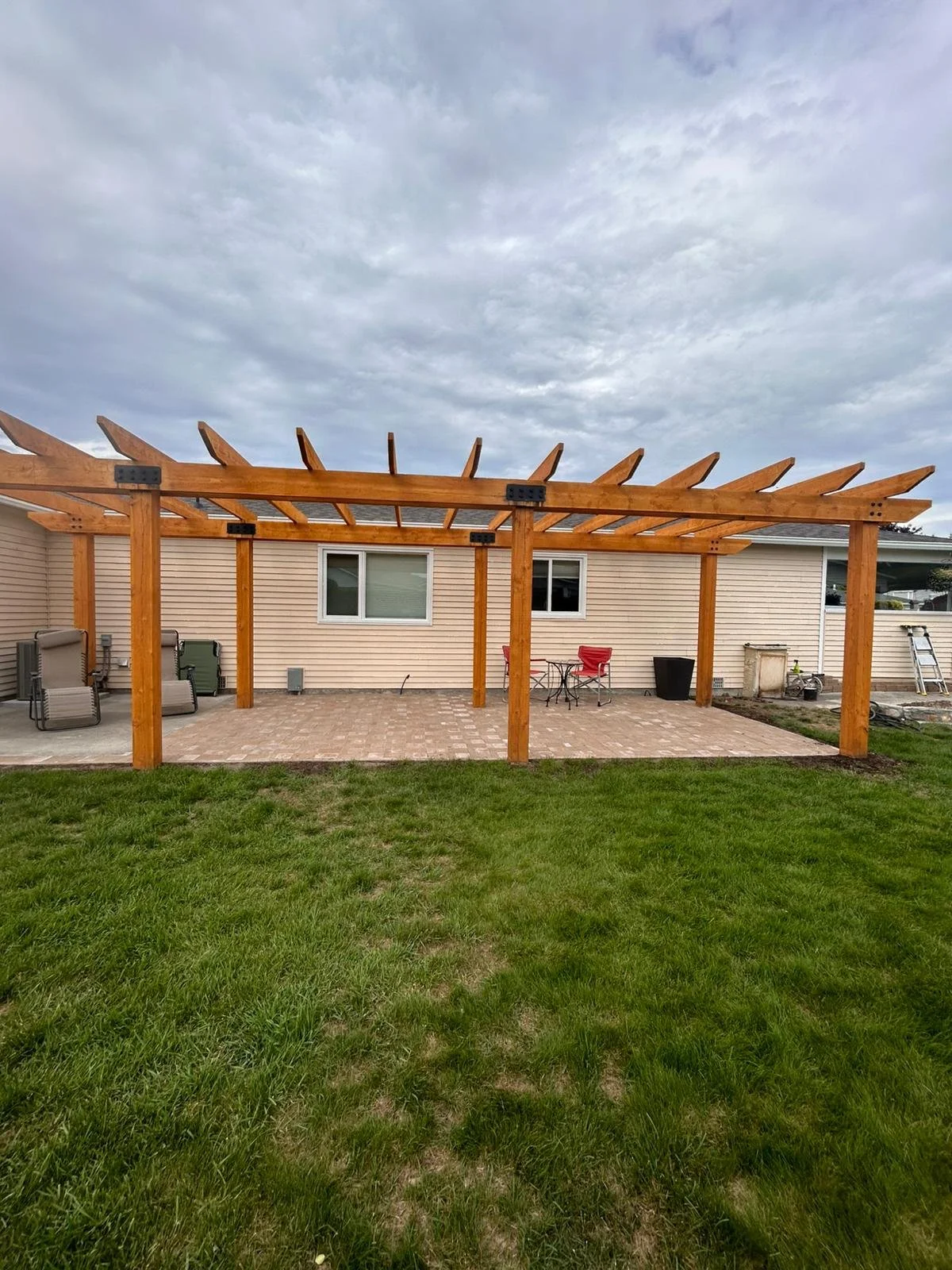 Newly built wooden pergola over a brick patio area behind a beige house, with chairs, a table, and outdoor equipment on the patio.