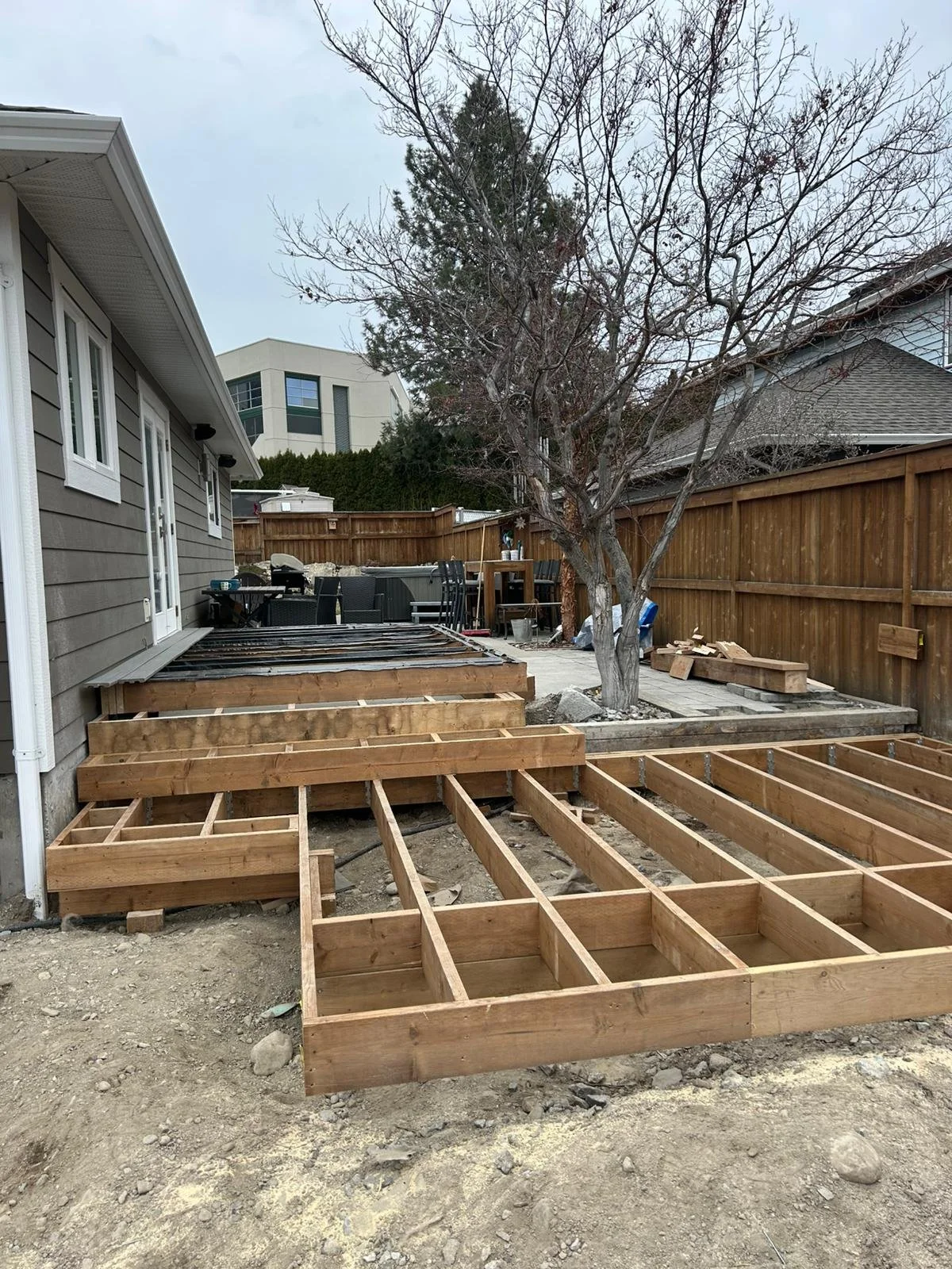 Wooden framework for a deck construction in a backyard with a tree, patio furniture, and a neighboring house in the background.