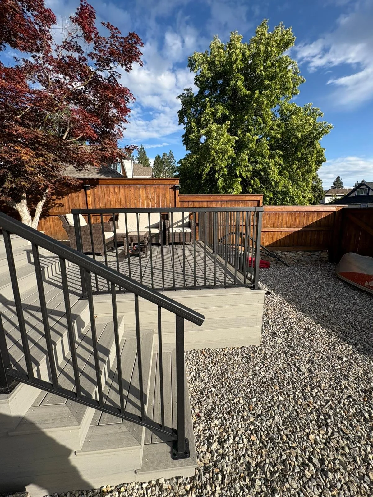 Backyard deck with outdoor furniture, a wooden privacy fence, trees, and a gravel ground surface under a partly cloudy sky.