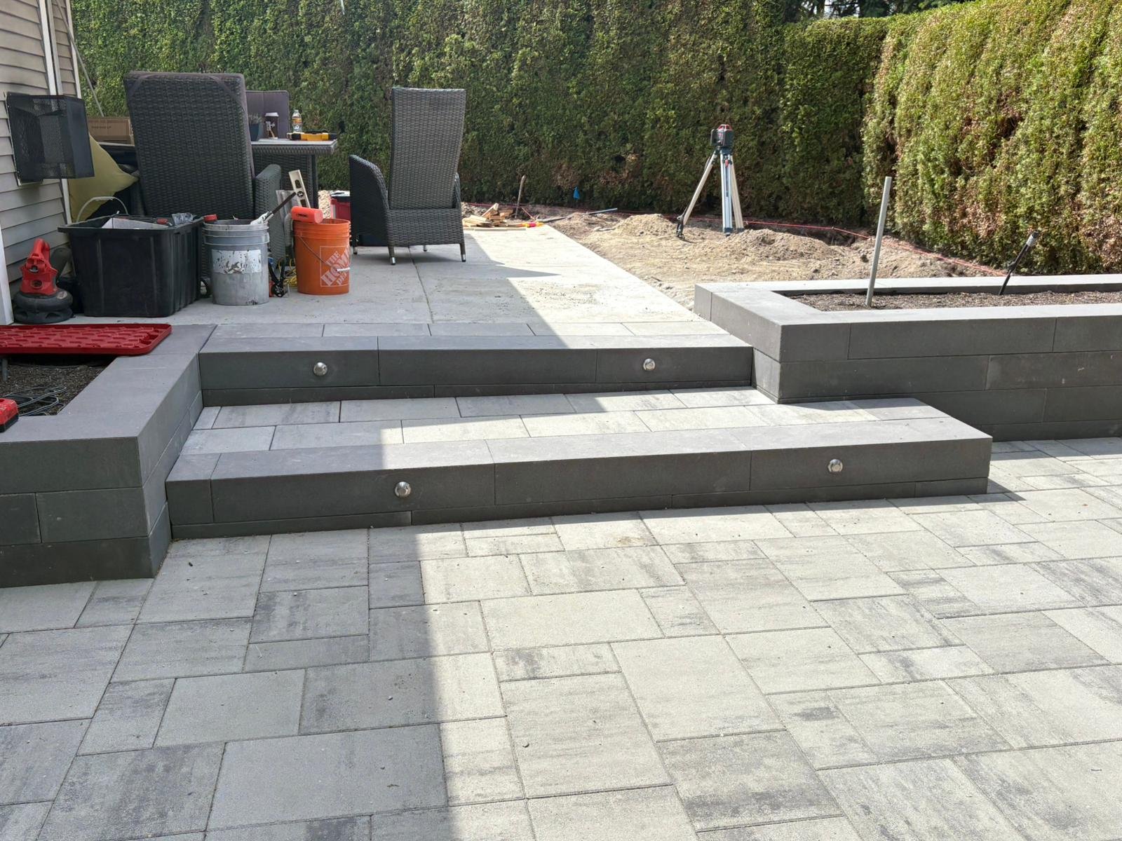 A partially completed outdoor patio with gray tile steps, seating area with black wicker chairs, and garden bed, surrounded by a green hedge.