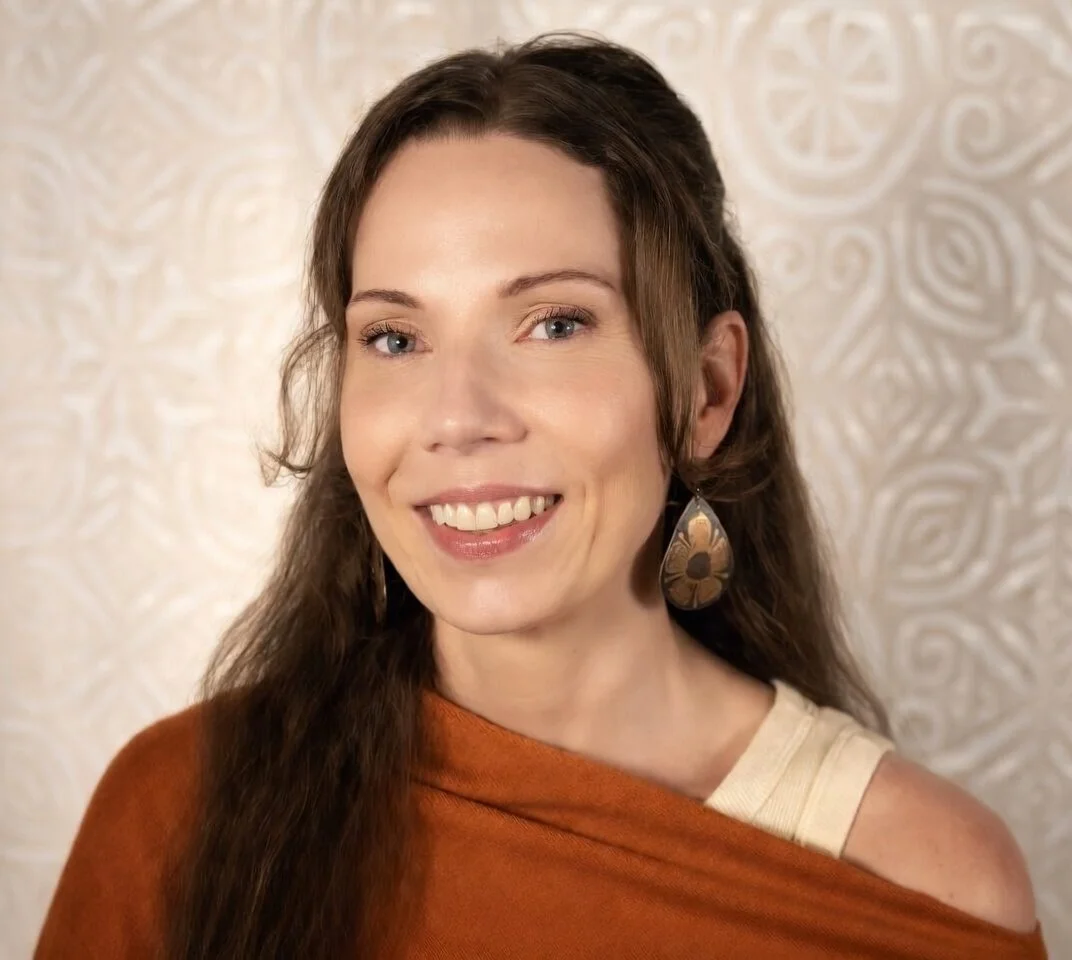 A woman with long brown hair wearing a beige top and a rust-colored shawl, smiling, with large dangling earrings, standing against a patterned cream-colored background.