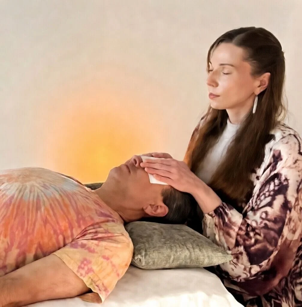 A woman with closed eyes gently placing her hands on a man's forehead while he is lying down with his eyes closed, on a pillow.