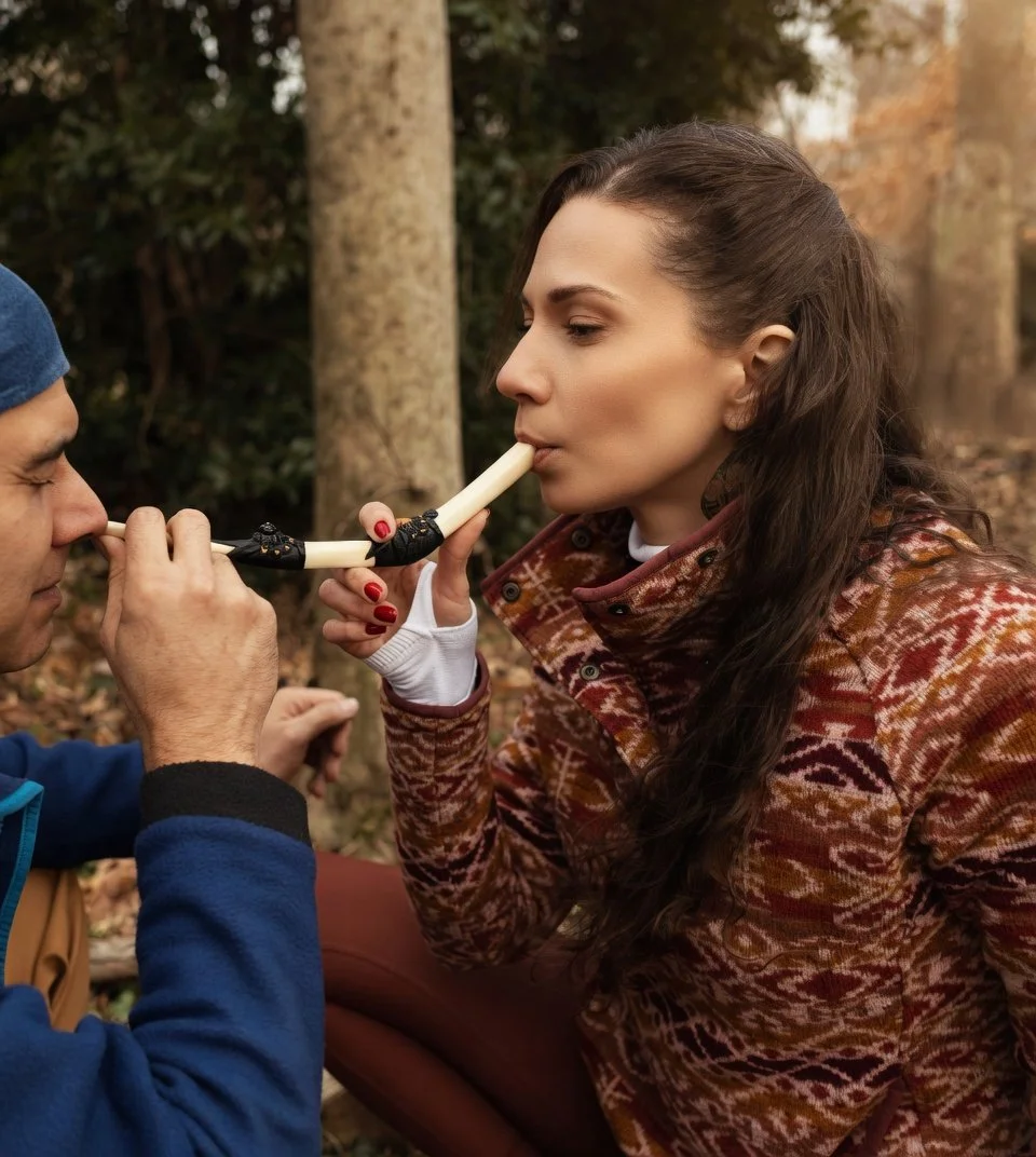 A woman is holding a long, white, curved object near her mouth while a man holds the other end towards his nose in a forest setting.