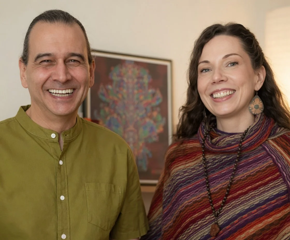 A smiling man in a green shirt and a smiling woman in a colorful shawl with earrings and a necklace, standing in front of a framed colorful artwork on a beige wall.