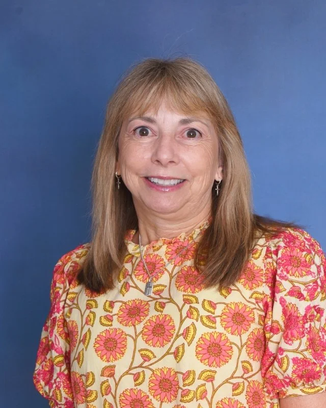 A woman with shoulder-length brown hair, wearing a blue floral-patterned blouse and coral-colored pants, smiling at the camera against a plain light background.