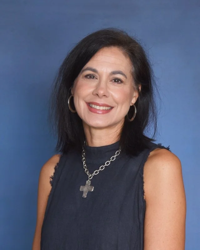 A woman with shoulder-length black hair, smiling, wearing a cream-colored sleeveless top, a pearl and gold chain necklace, matching earrings, and a gold bracelet, seated against a plain light background.