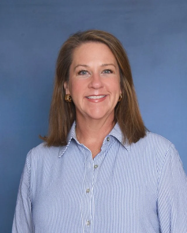 A woman with shoulder-length brown hair, smiling, sitting with hands clasped, wearing a white short-sleeve blouse and a silver watch, against a plain light background.