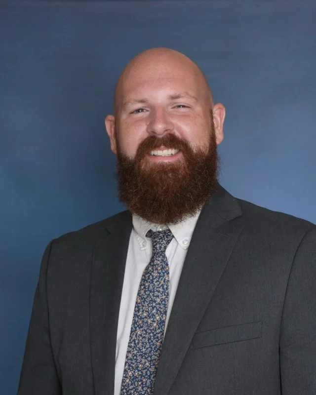 A man with a bald head and a full beard, wearing a dark blazer, a striped shirt, and a patterned tie, sitting against a plain light background, smiling at the camera.