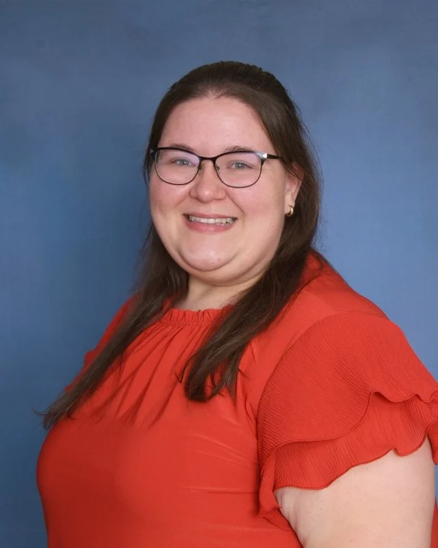 A woman with long brown hair, glasses, and gold earrings, smiling in a burgundy dress with ruffled sleeves and a dotted pattern, standing against a plain light background.