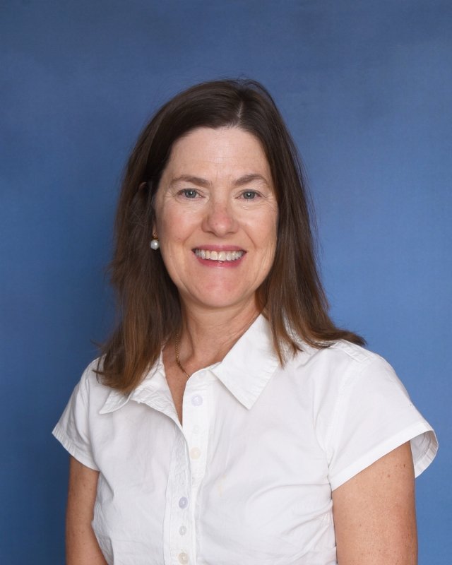 A smiling woman with shoulder-length brown hair wearing a colorful floral shirt with pink, red, orange, green, blue, and purple patterns. She has small hoop earrings, a gold necklace, and a black watch, standing against a plain white background.