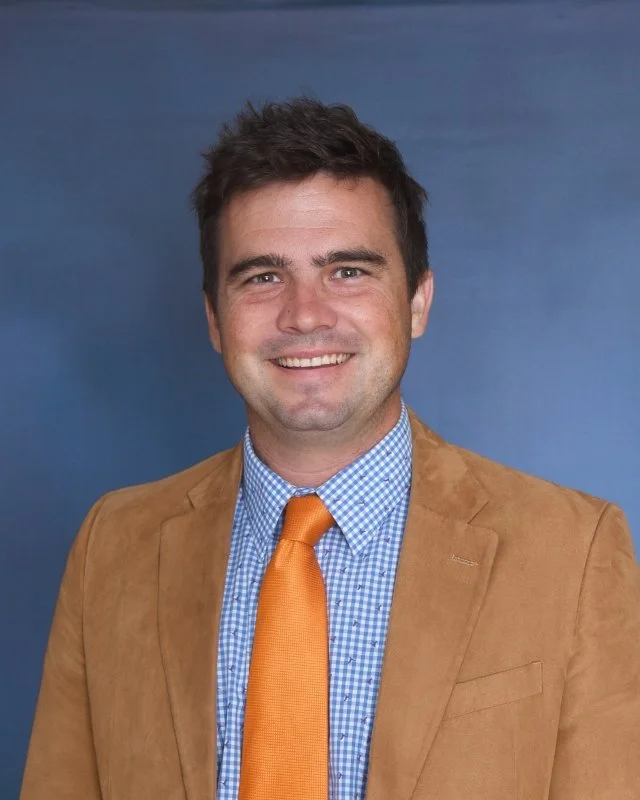 A man in a gray suit with a rust-colored shirt and a navy tie with red dots, sitting against a plain light background.