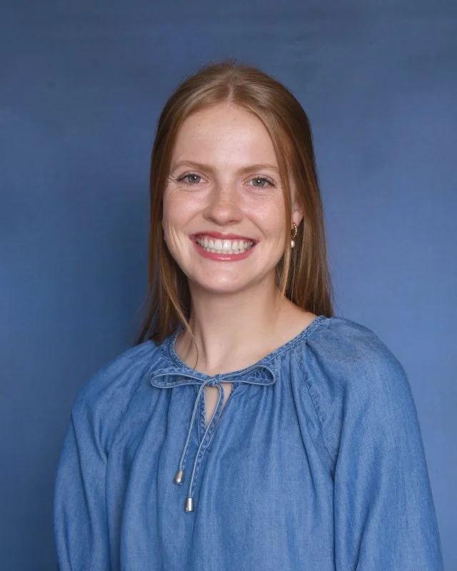 A smiling woman with long red hair wearing a colorful patchwork-style blouse with a tie-neck, seated against a light gray background.