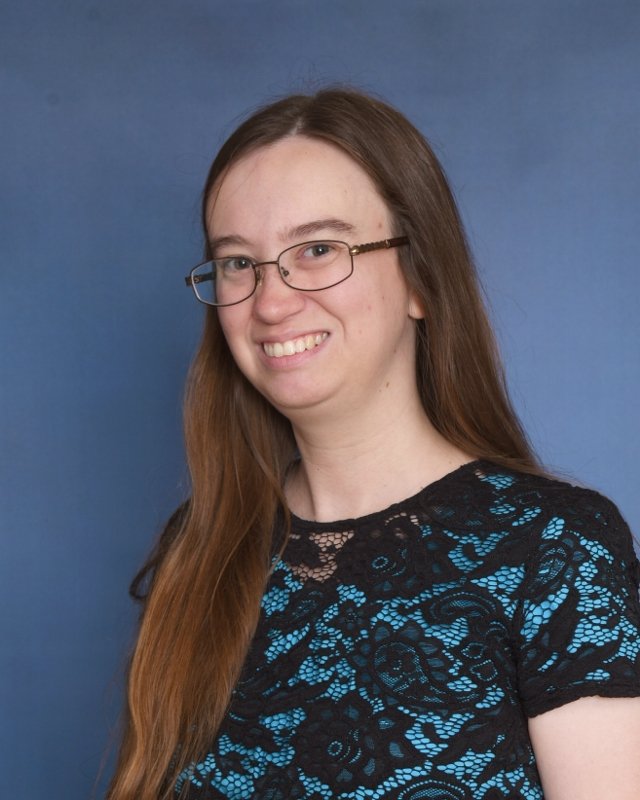 A woman with long brown hair, glasses, and a patterned dress, smiling at the camera against a plain light background.