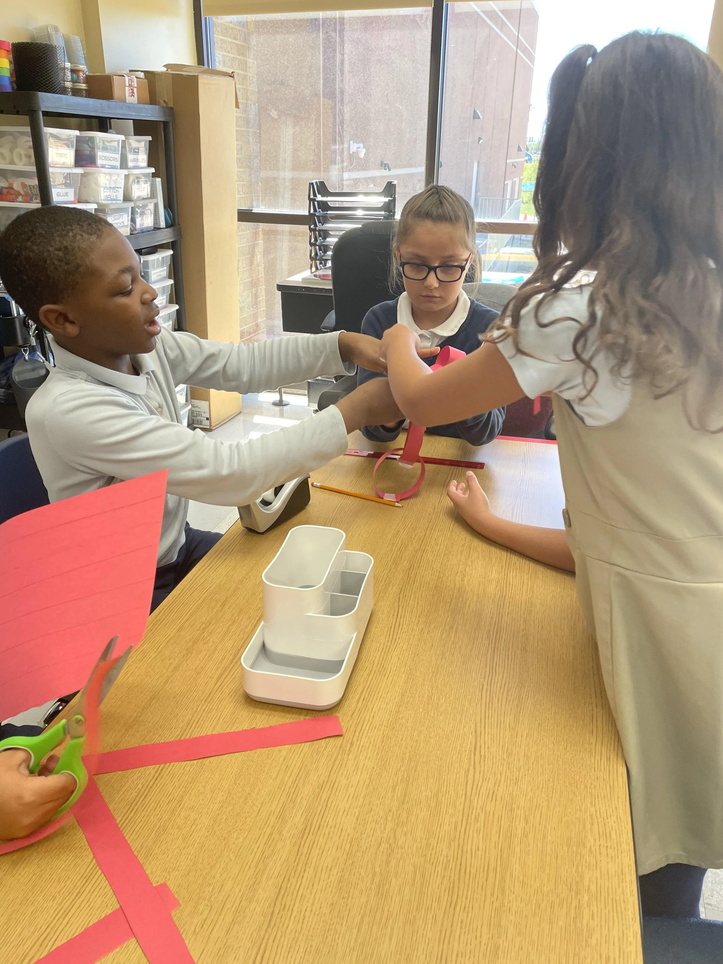 Three children working together at a table, with one child passing an item to another, surrounded by school and craft supplies.