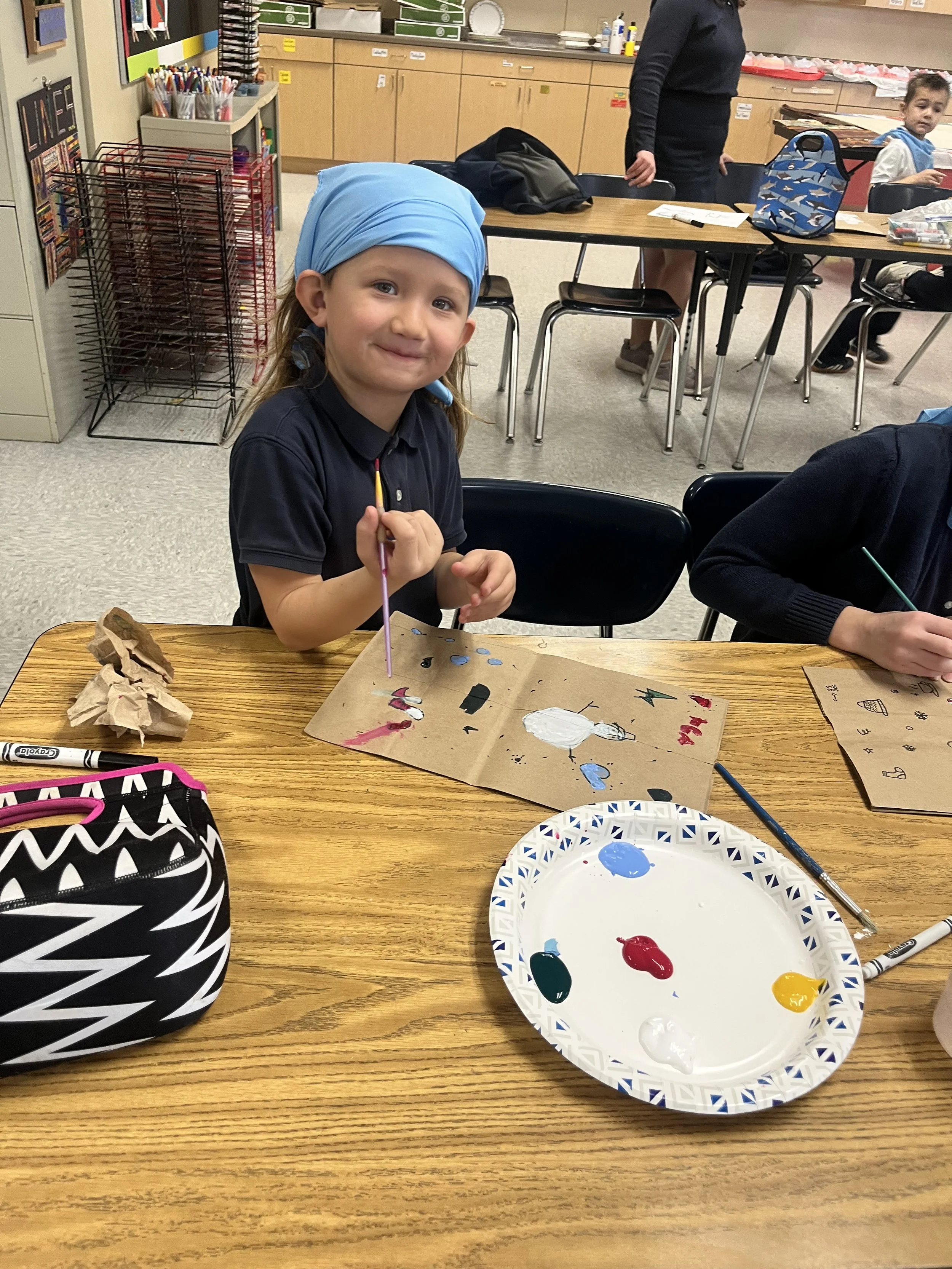 A young girl in a black shirt and blue headscarf sitting at a table with art supplies, smiling while holding a paintbrush. There is a paper plate with paint colors and a paper with painted artwork in front of her. The classroom background includes other students, tables, chairs, and classroom supplies.