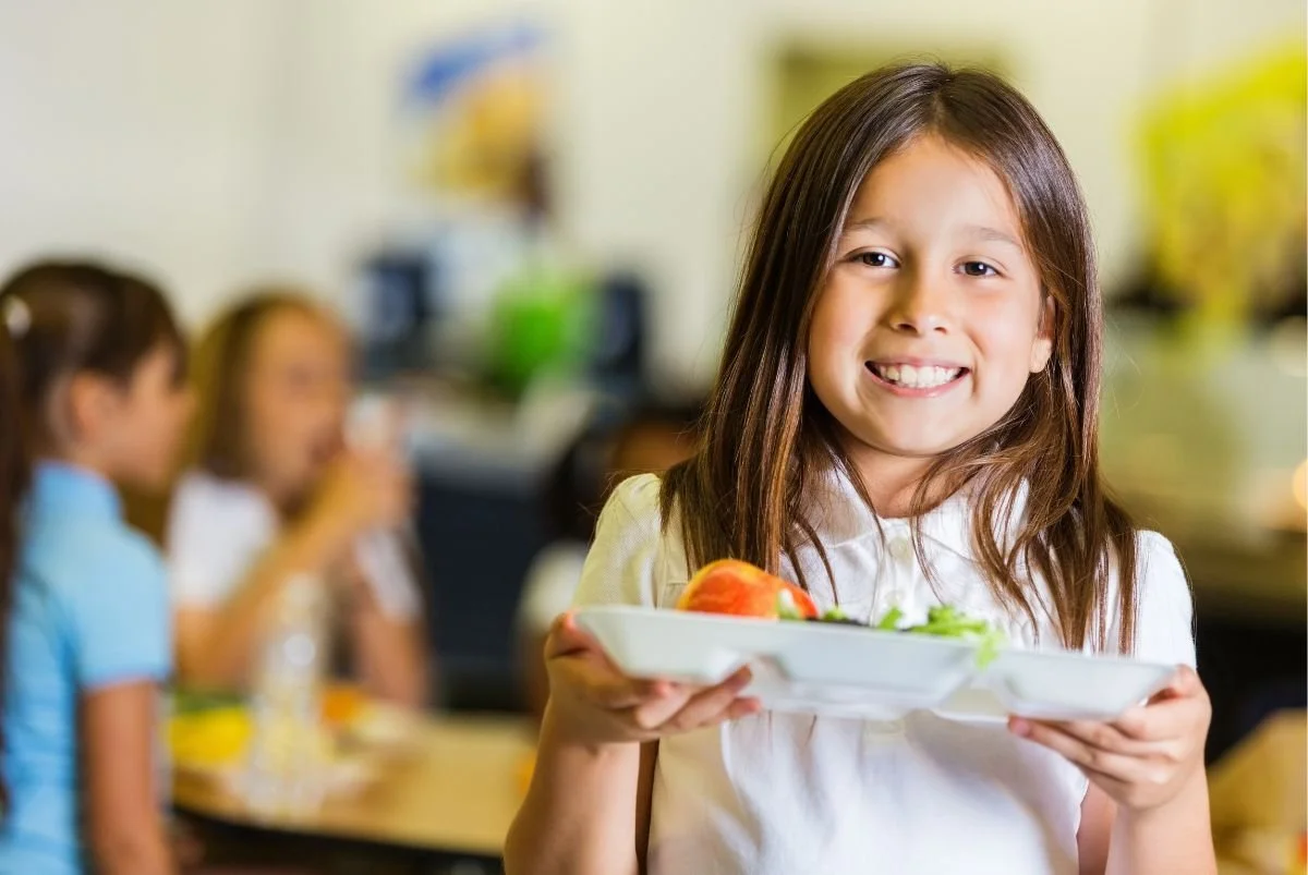 A young girl smiling and holding a plate of food in a school cafeteria, with other children blurred in the background.
