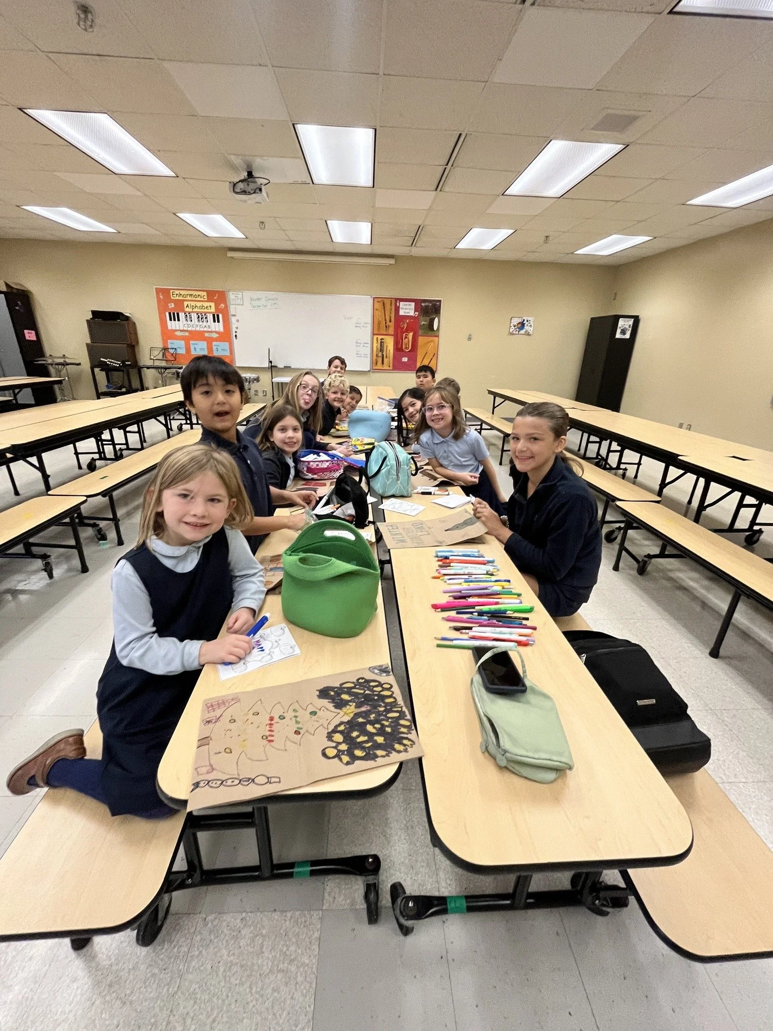 School classroom with young students sitting at tables, working on arts and crafts projects with markers, paper, and backpacks.