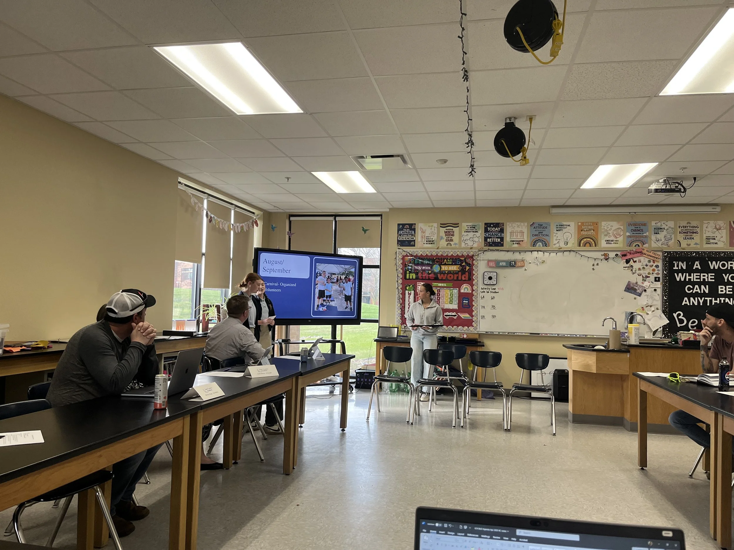 Classroom with students and teachers during presentation, large display screen showing August/September activities, whiteboard, chairs, desks, posters, and decorations on the walls.