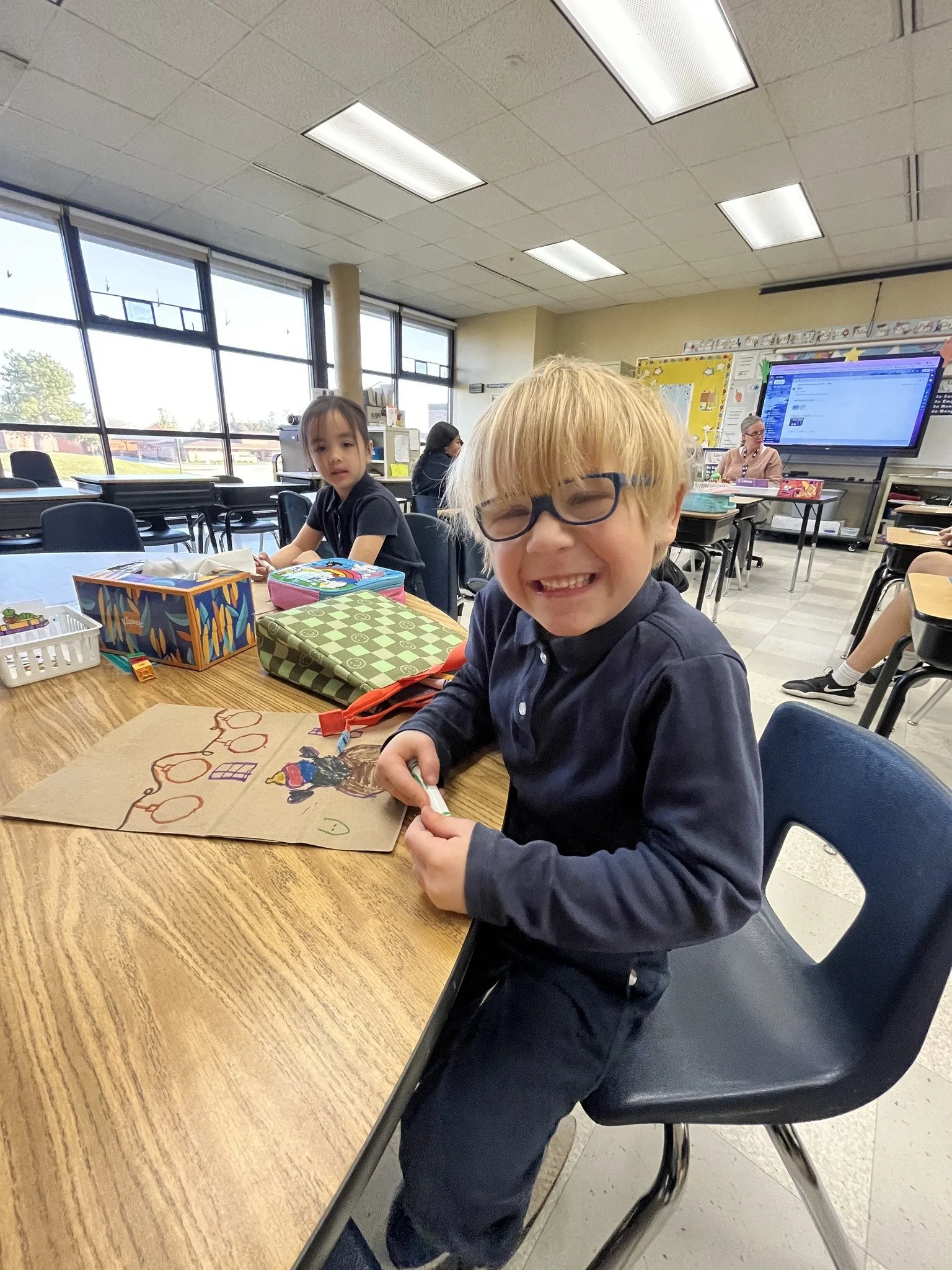 A young boy with blonde hair, glasses, and a wide smile sitting at a school desk with a Valentine's Day drawing. Other children and a teacher are visible in the background in a classroom with large windows and a digital whiteboard.