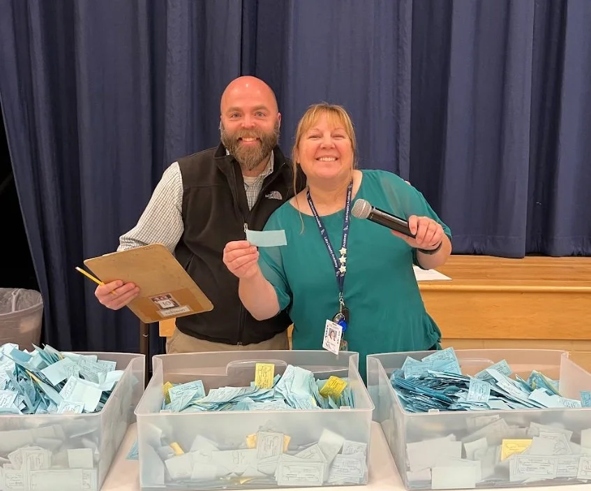 Two smiling election workers standing behind tables filled with ballots, one woman holding a microphone and a piece of paper, a man holding a clipboard, with a blue curtain in the background.
