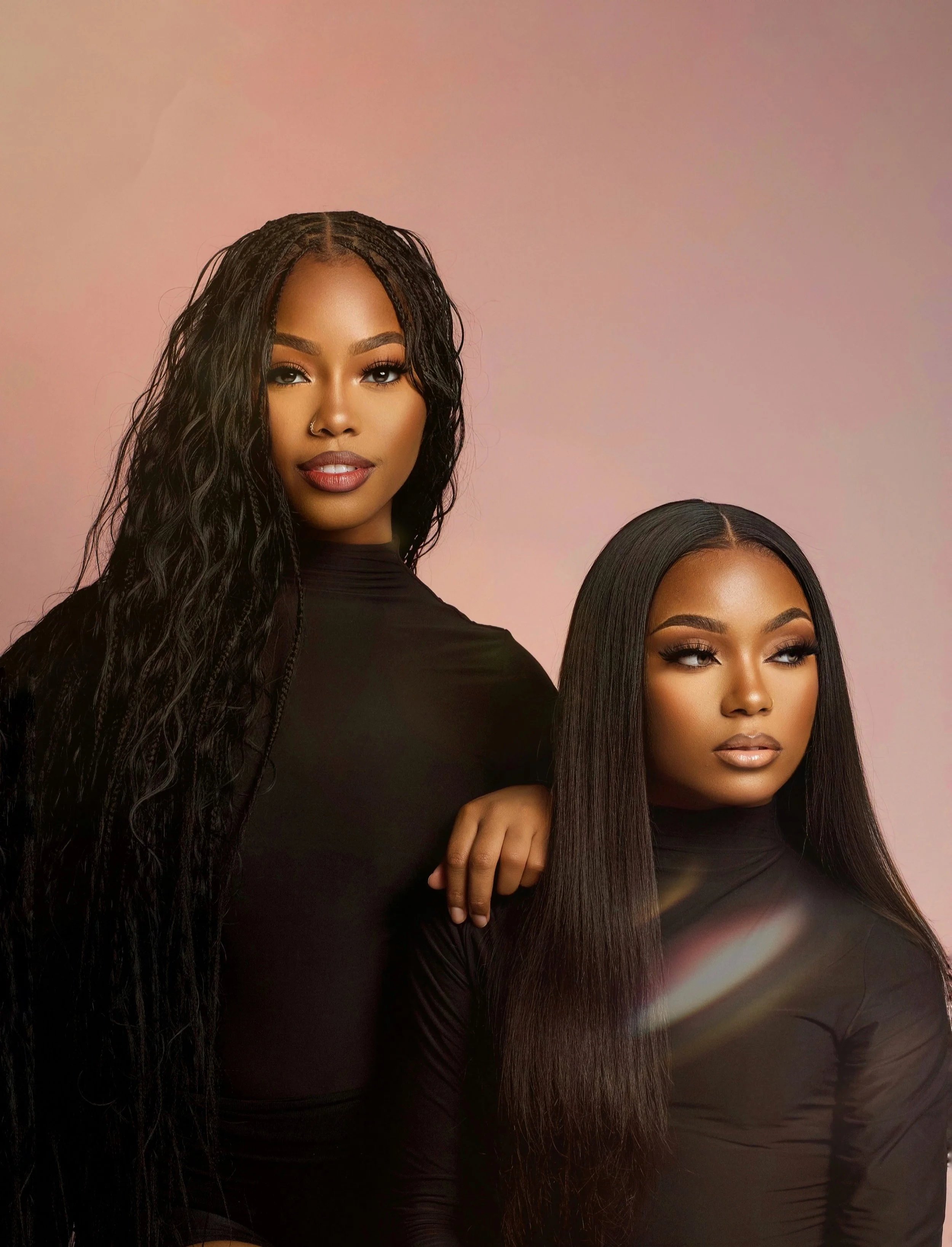 Two women with long straight black hair wearing black outfits posing against a pink gradient background.