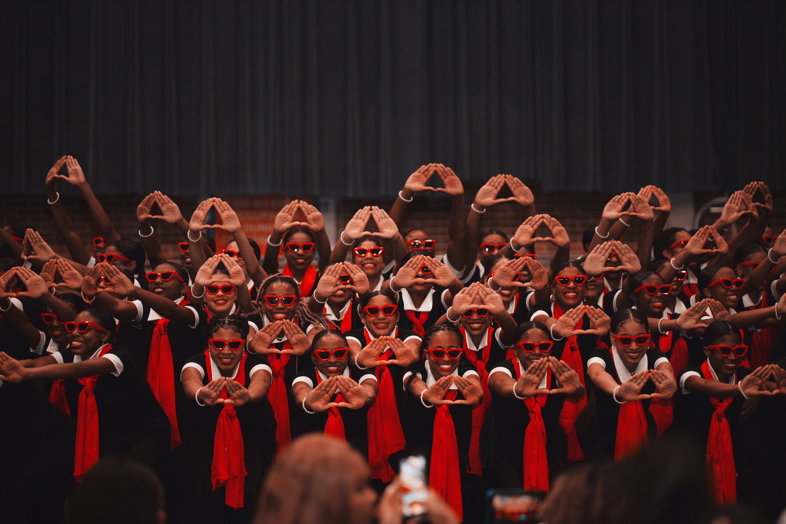 Group of performers in black costumes with red scarves, red sunglasses, and white pearl jewelry, dancing on stage with a dark backdrop.