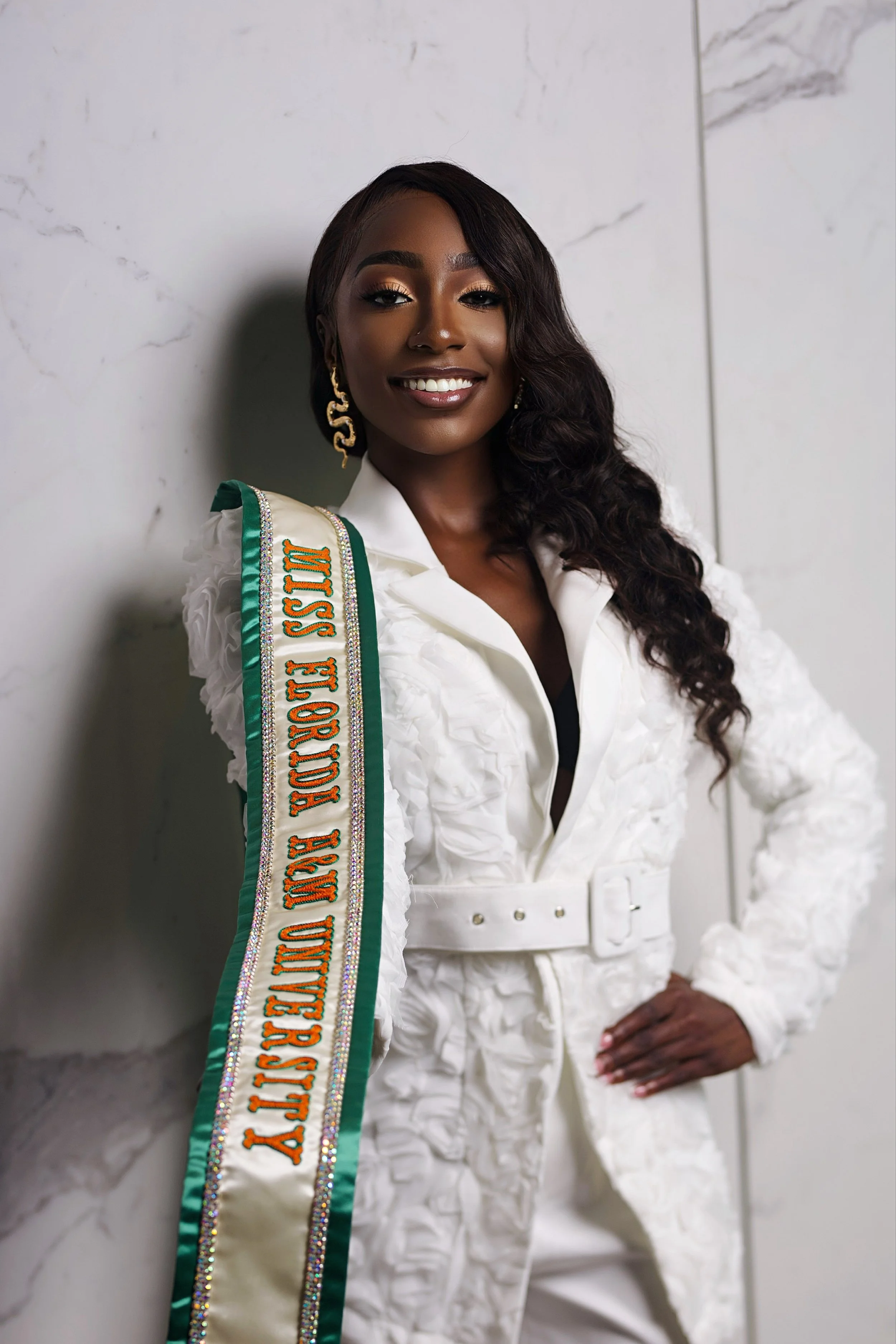 A woman wearing a white textured blazer with a matching belt, gold earrings, and a sash that reads "Miss Texas USA." She has long, wavy dark hair and is smiling while standing against a marble wall.