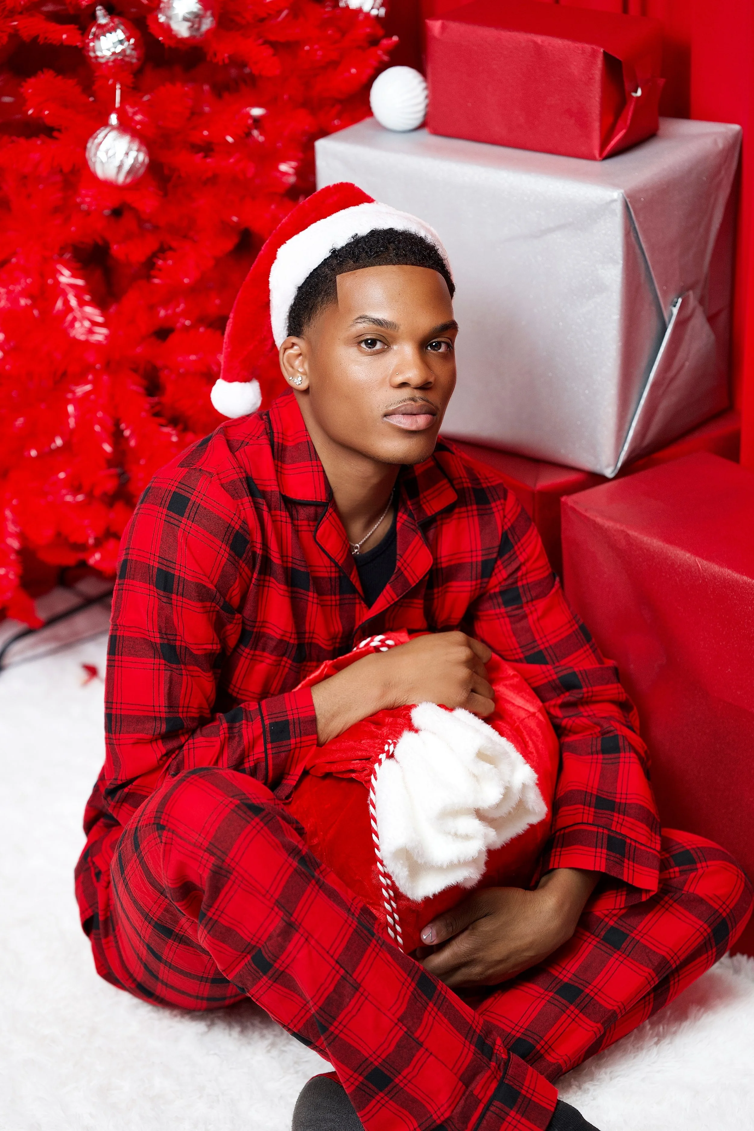 A young man in red plaid pajamas and a Santa hat sitting on the floor, holding a Christmas stocking in front of a decorated Christmas tree with wrapped presents nearby.