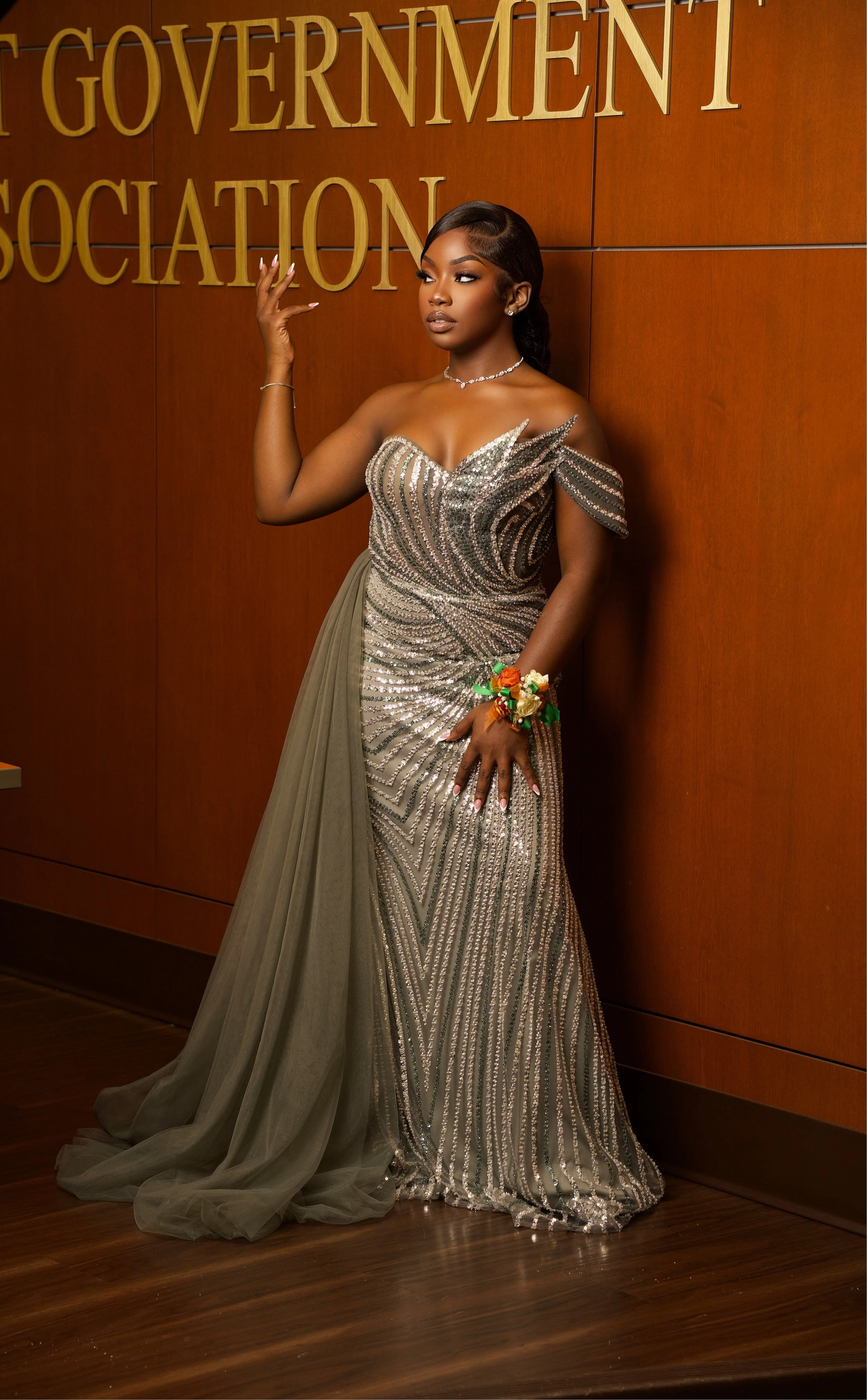 A woman in a glamorous, silver, sequined evening gown with a tulle train, standing in front of a wooden wall with gold letters says 'Government Association', wearing jewelry and a floral corsage, posing with one hand near her face.