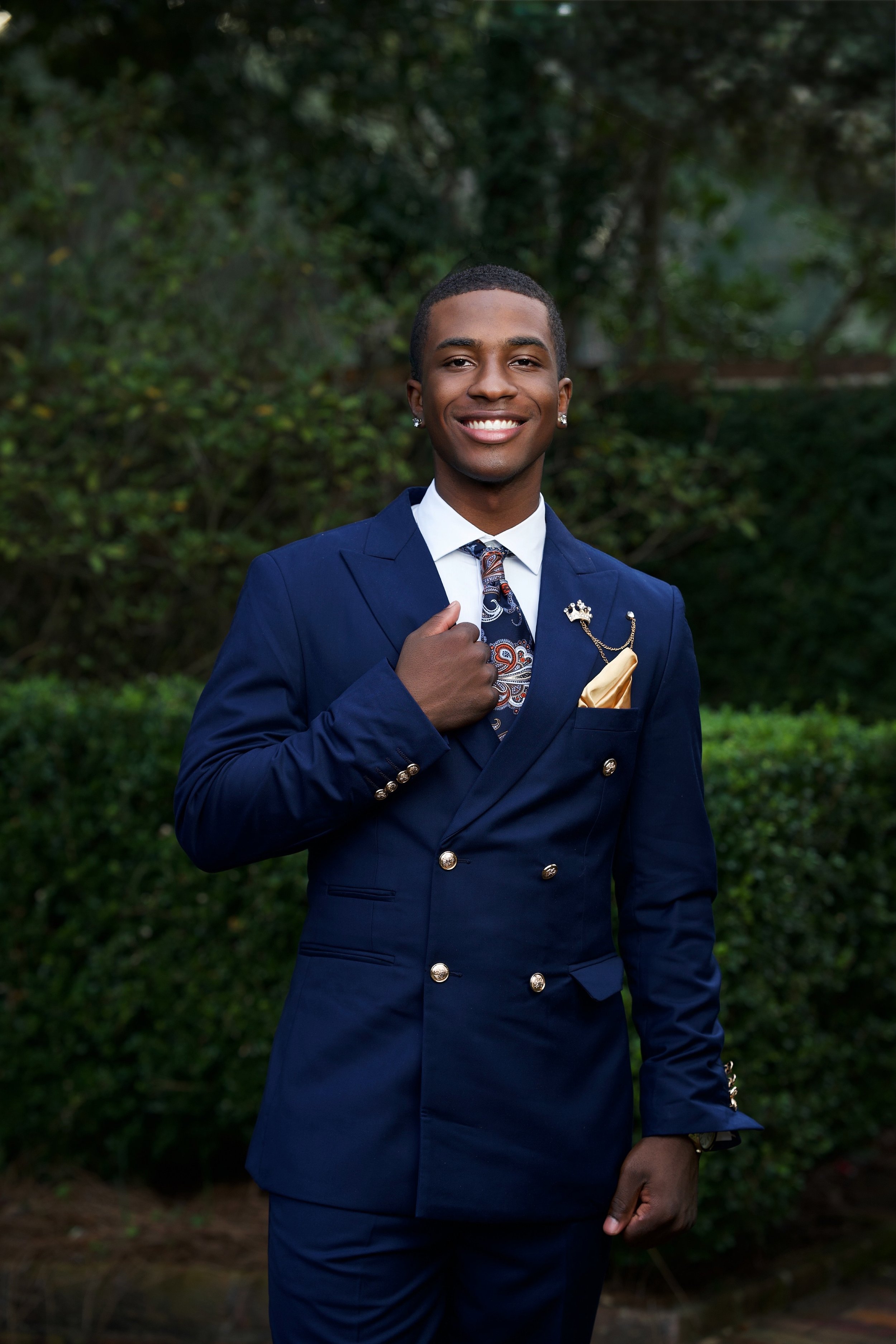 A young man in a navy blue double-breasted suit with gold buttons, white shirt, paisley tie, and yellow pocket square. He is smiling and standing outdoors with trees and bushes in the background.