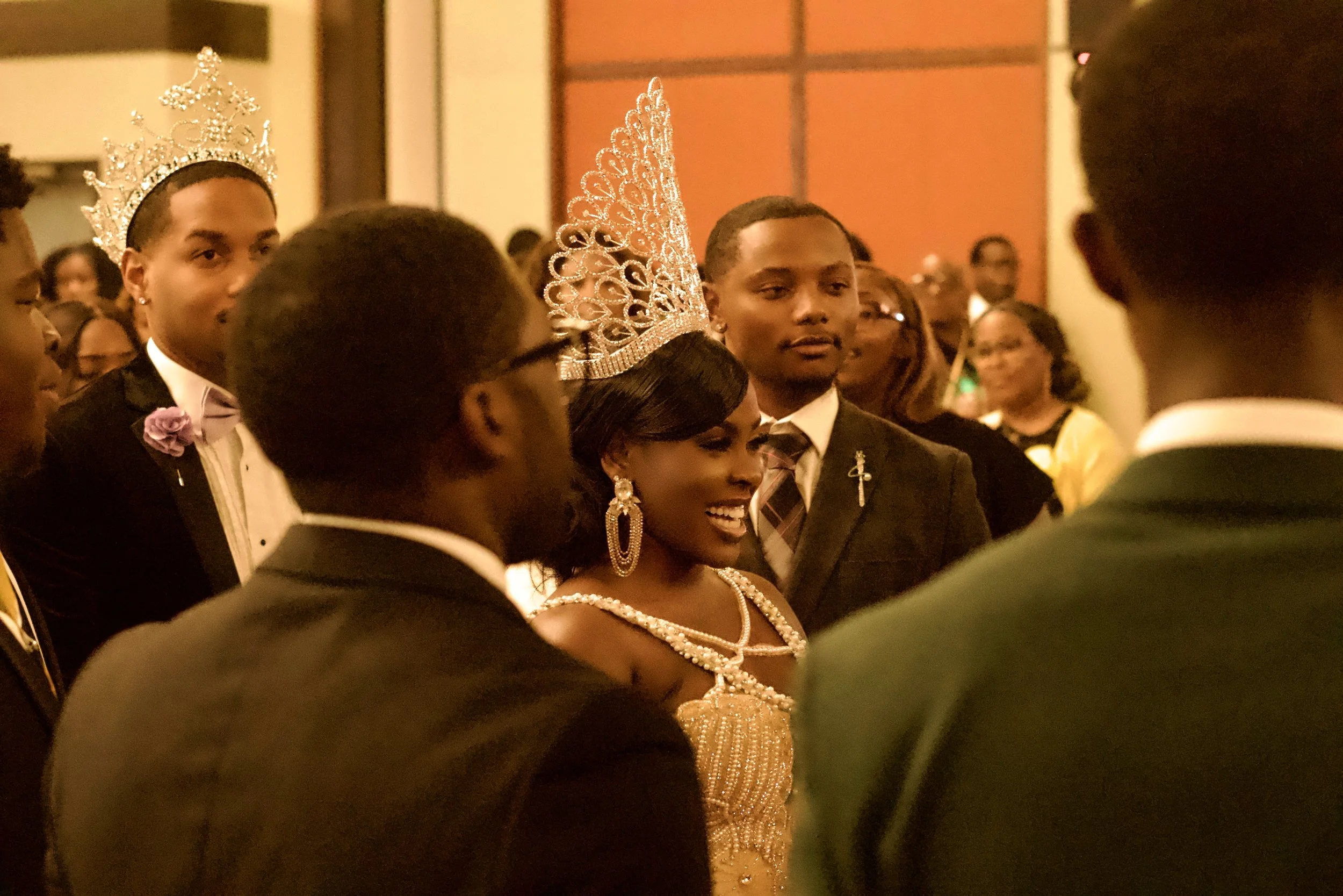 A woman wearing a crown and jewelry standing among men in suits at an event, with some people smiling in the background.