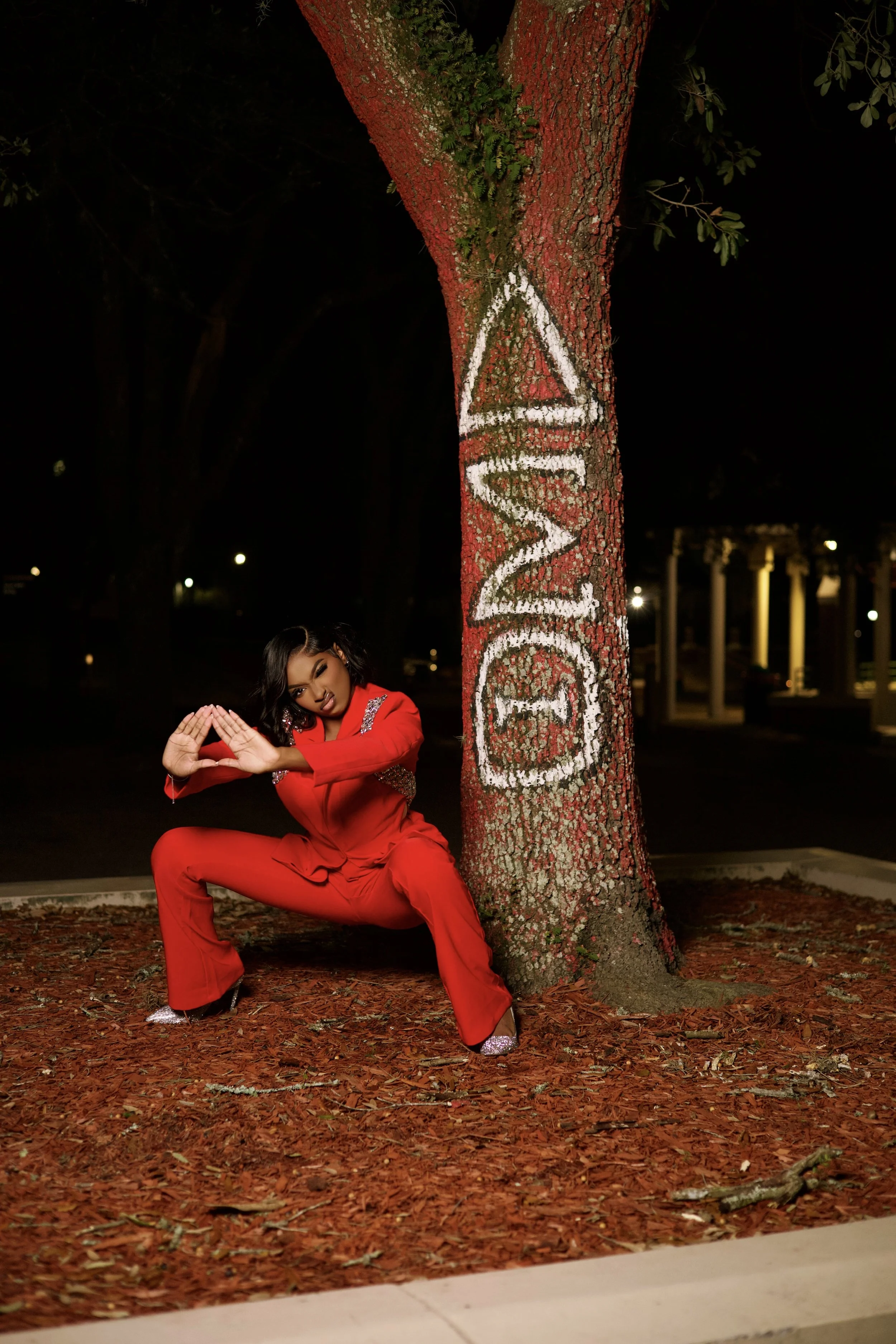 A woman in a red outfit squatting near a tree with the word 'LOVE' painted on the trunk and forming a heart shape with her hands, at night.